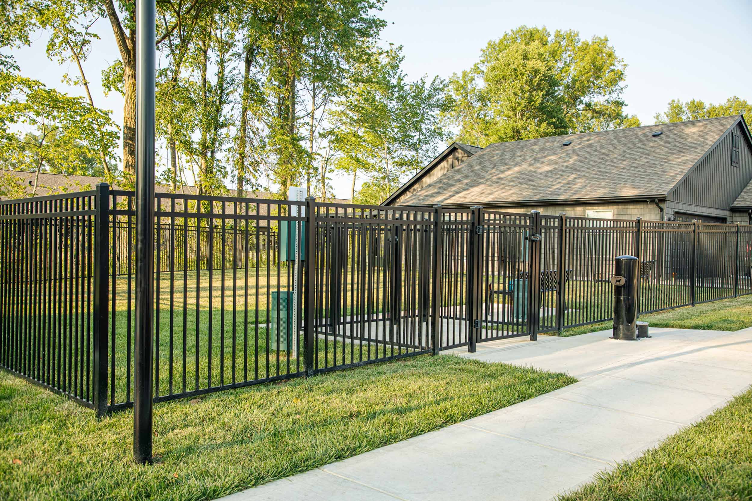 Black metal fence surrounding a grassy outdoor area with mature trees, a sidewalk, and a building with a sloped roof.