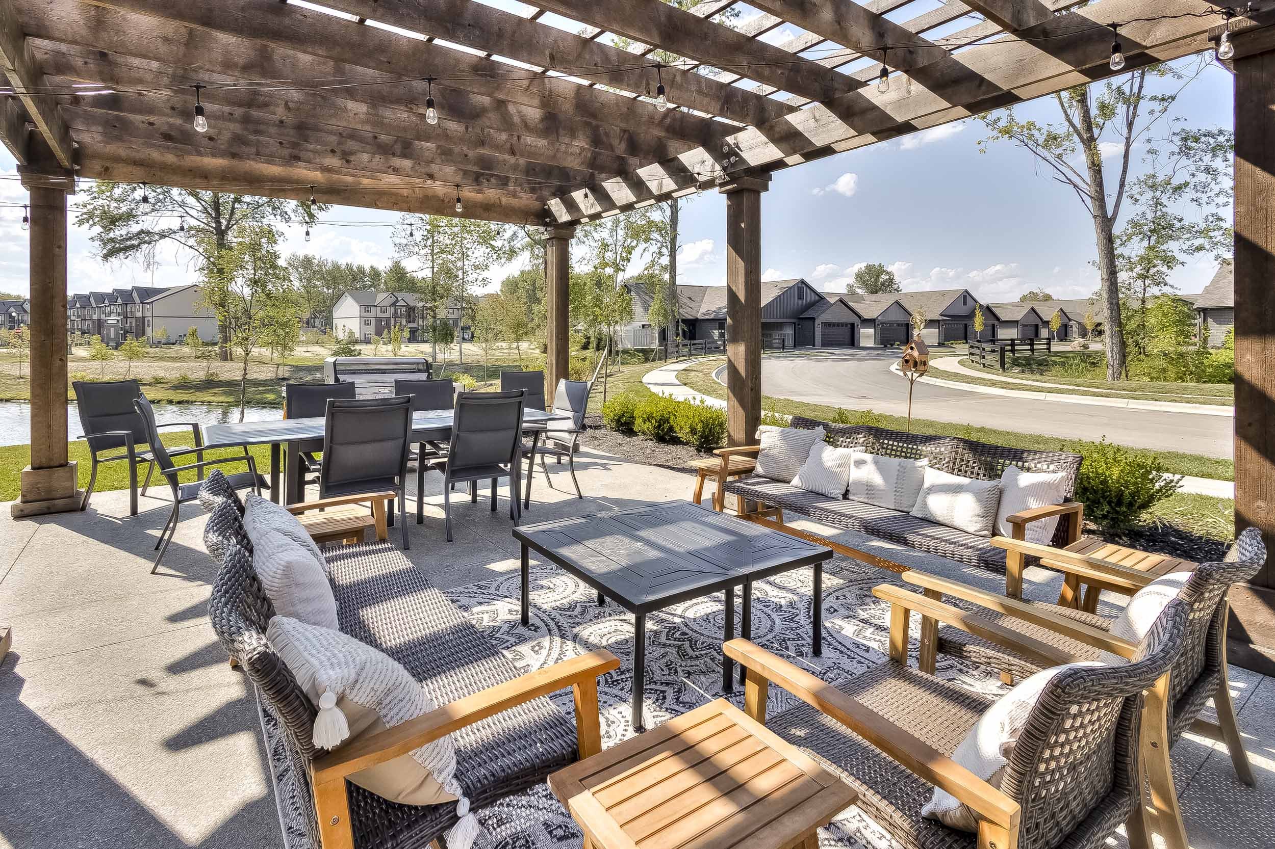 Outdoor patio area with a wooden pergola, outdoor furniture including a dining table with eight chairs, a wicker sofa with white cushions, and a coffee table, overlooking a pond and residential neighborhood.