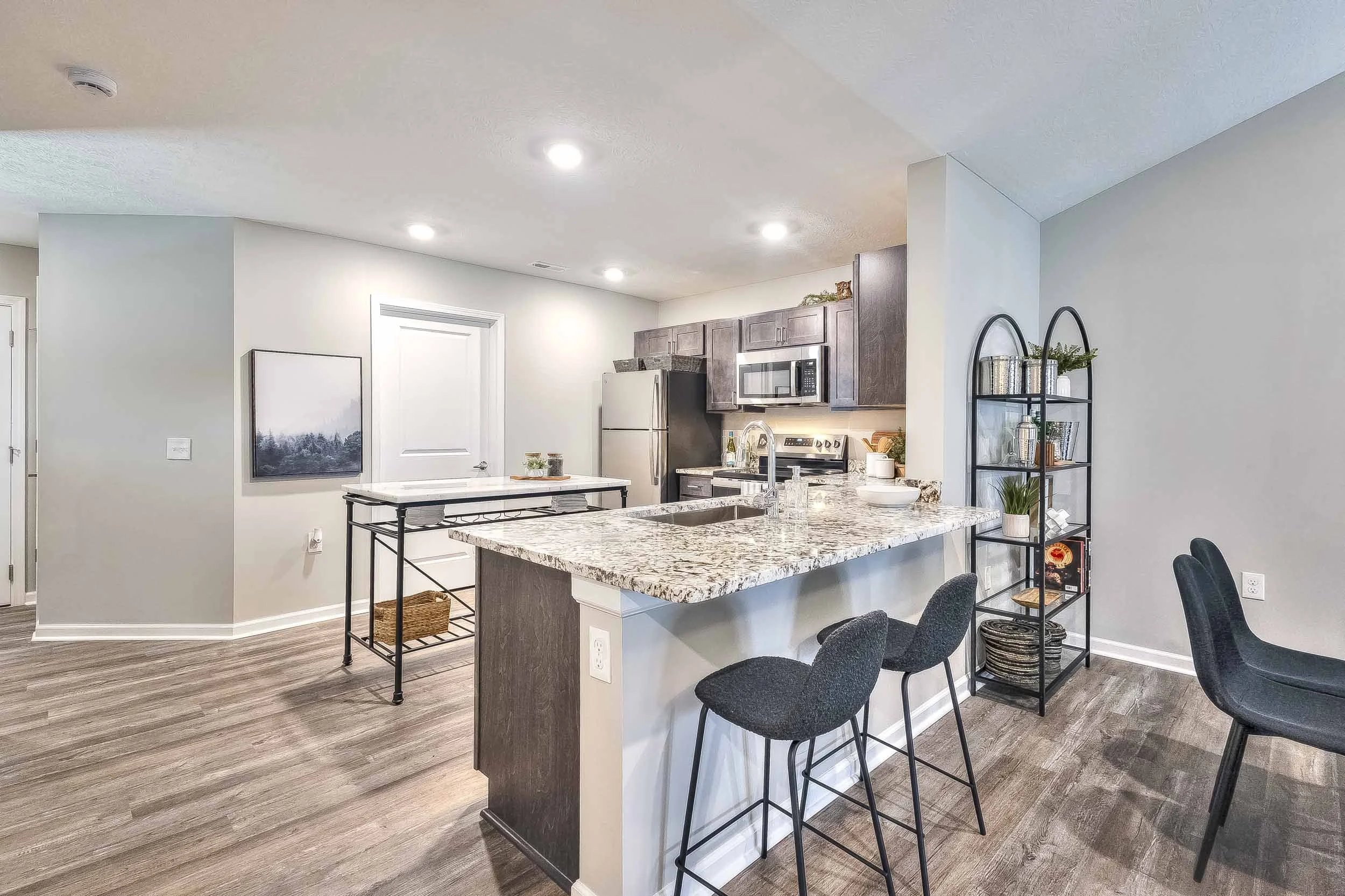 Modern kitchen with gray cabinets, granite countertops, stainless steel appliances including microwave, oven, and refrigerator, and a center island with seating, hardwood floors, and a black metal shelving unit.
