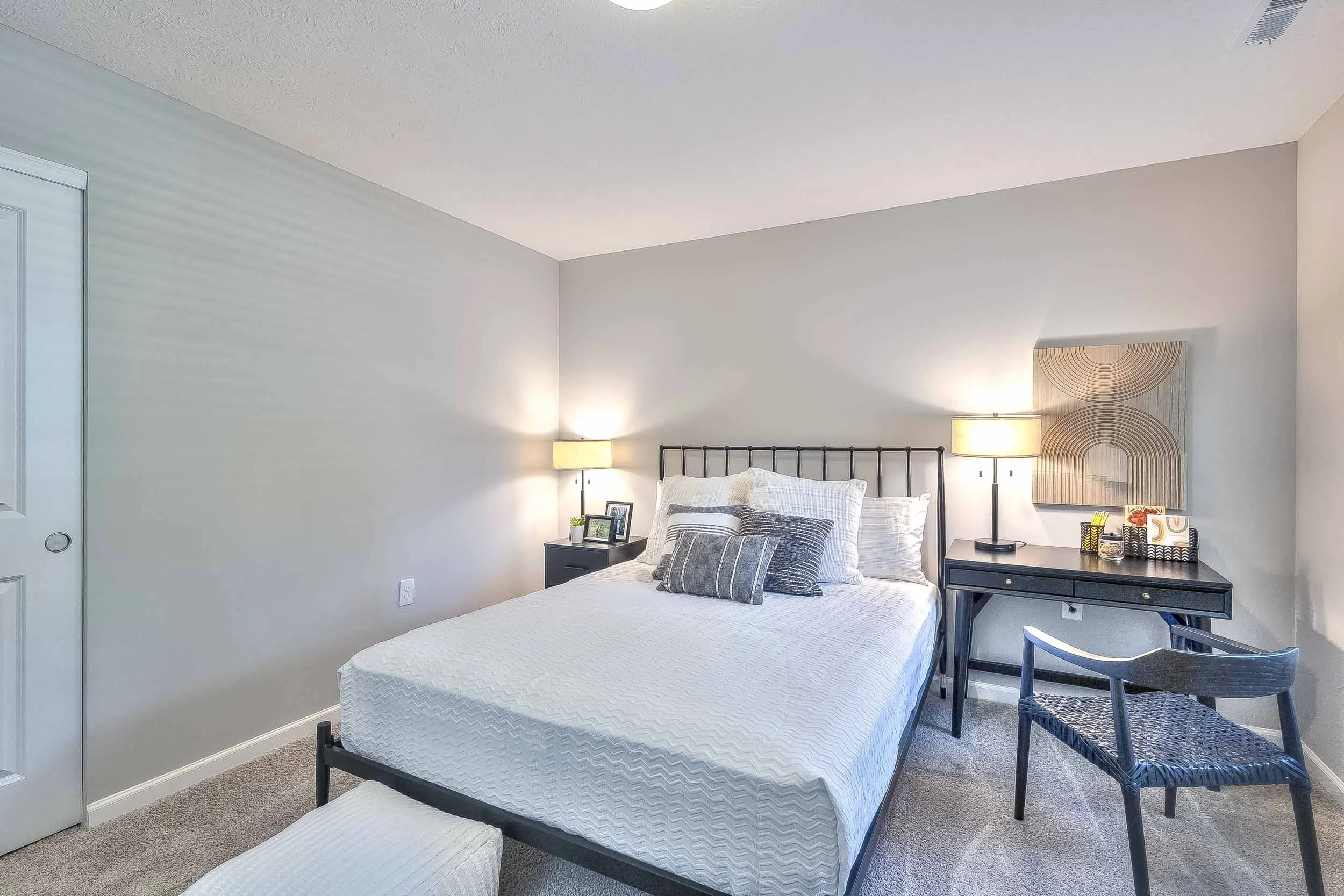 A neatly made bed with multiple pillows in a bedroom. There are two nightstands with lamps on each side, a chair and a small wooden table, and some decorations on the walls.