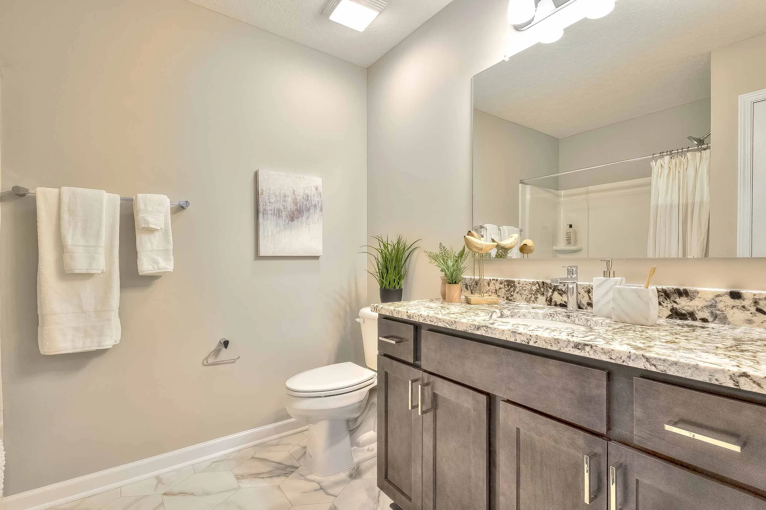 Modern bathroom with a granite countertop, wooden vanity, potted plants, large mirror, toilet, towel rack with towels, and shower with curtain