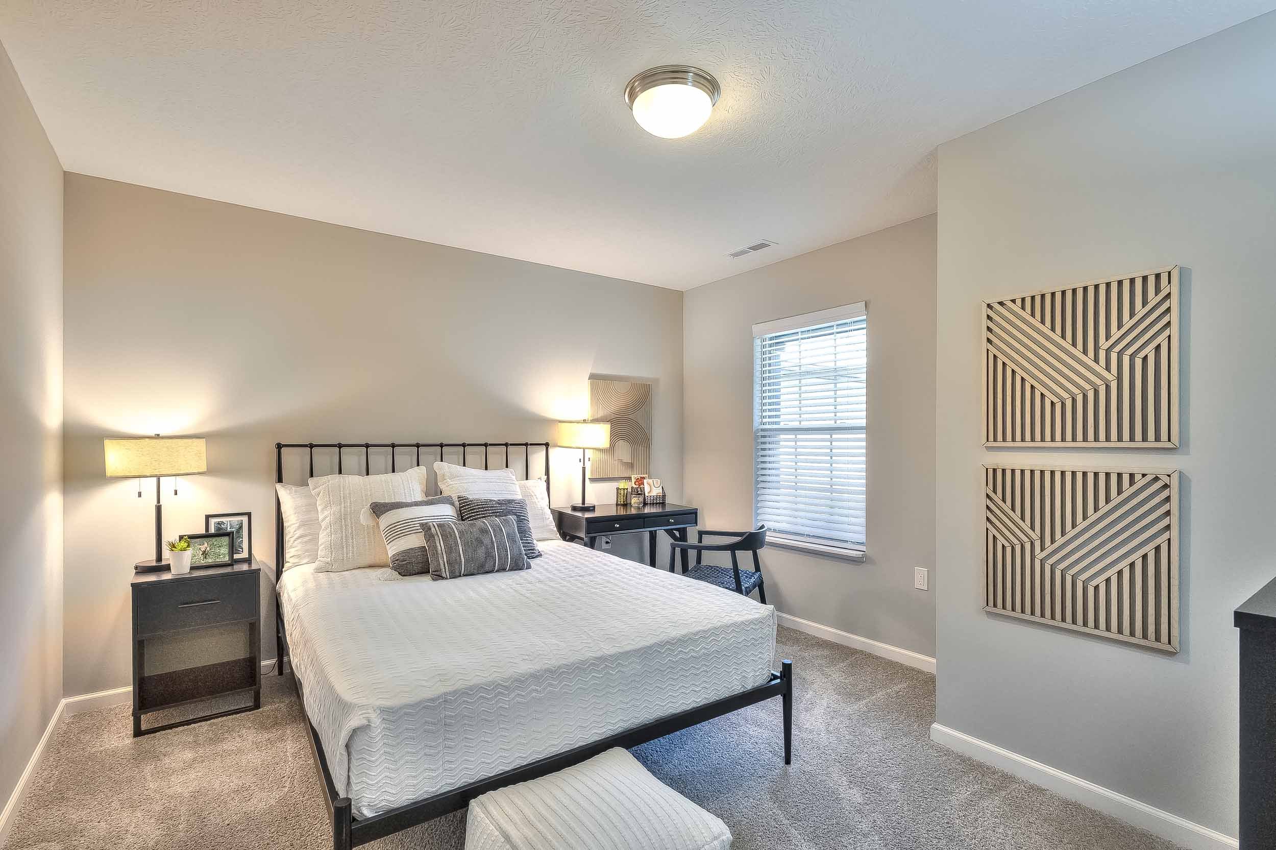 A bedroom with a bed, nightstand, desk, and window, decorated in neutral tones.