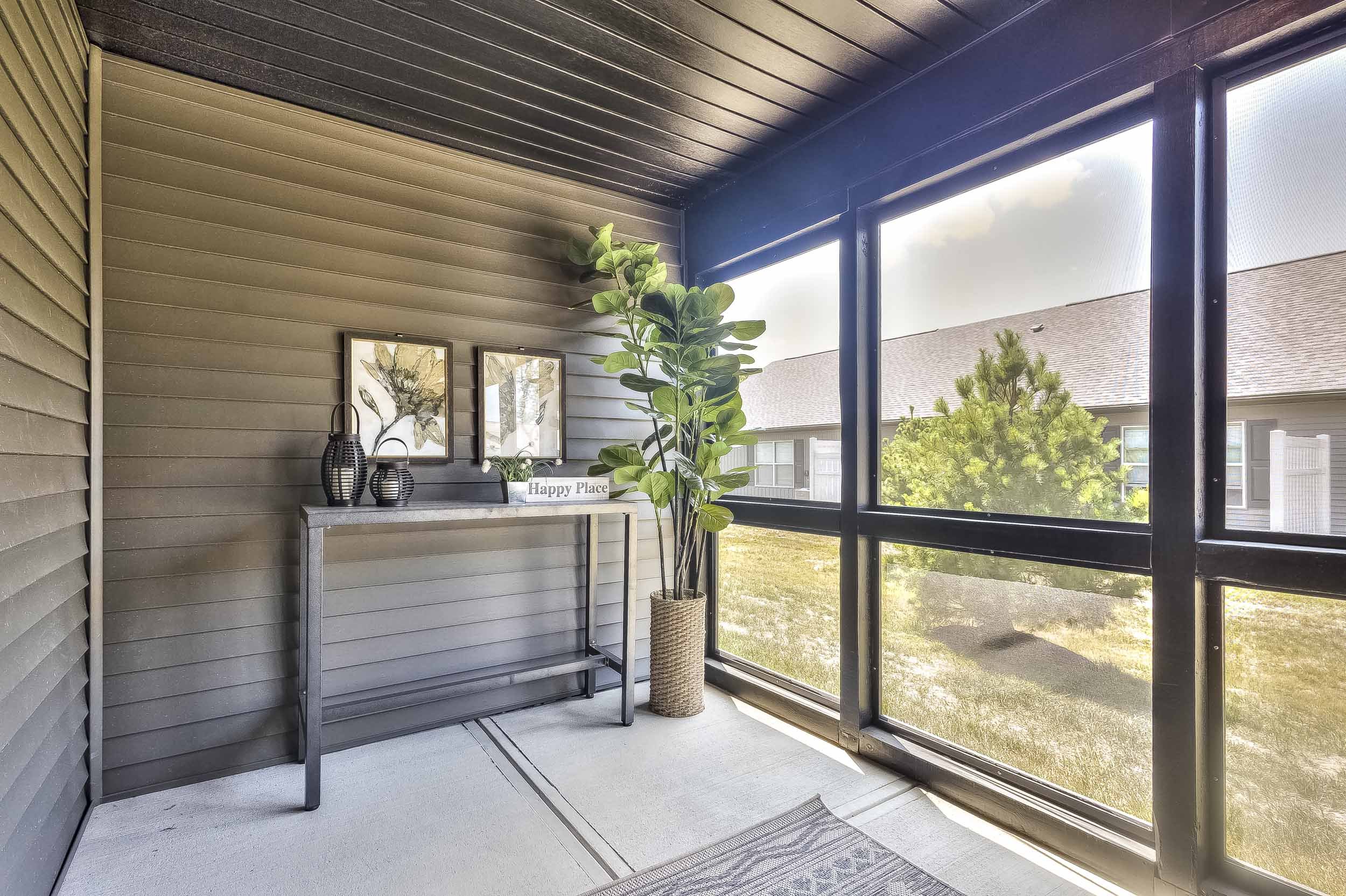 Enclosed balcony with dark wood ceiling, gray siding walls, large potted plant, and a narrow console table with decorative vases and framed leaf art, overlooking a yard with grass and trees.