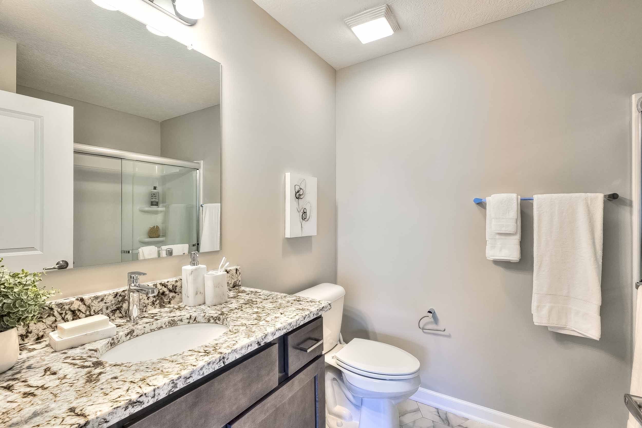 Modern bathroom with granite countertop, white sink, and a large mirror above. Toilet is next to the counter, with a white towel hanging on a bar on the wall. The room has white walls, a hexagonal tiled floor, and a bathroom shower with glass doors v