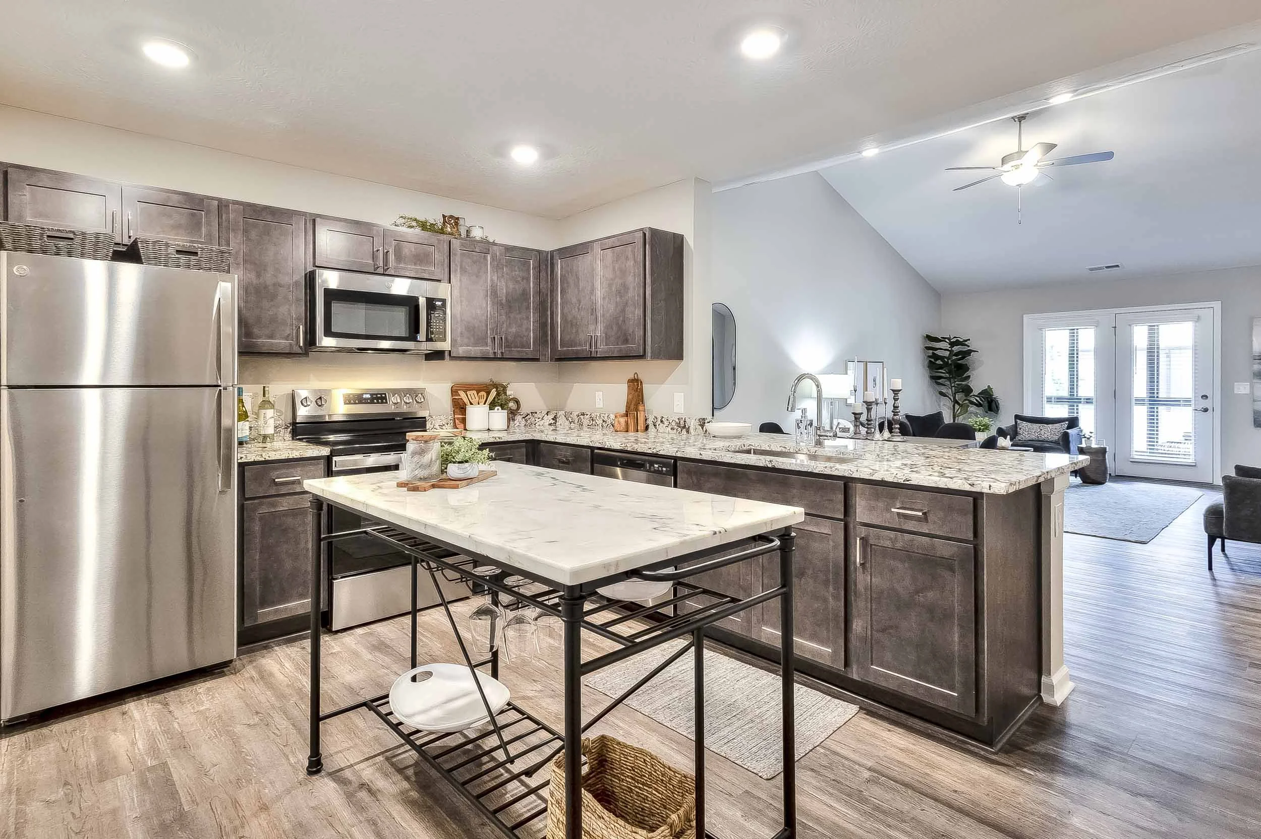 Modern kitchen with gray cabinets, stainless steel refrigerator, microwave, and oven, island with marble countertop, and an open living area with door to balcony.