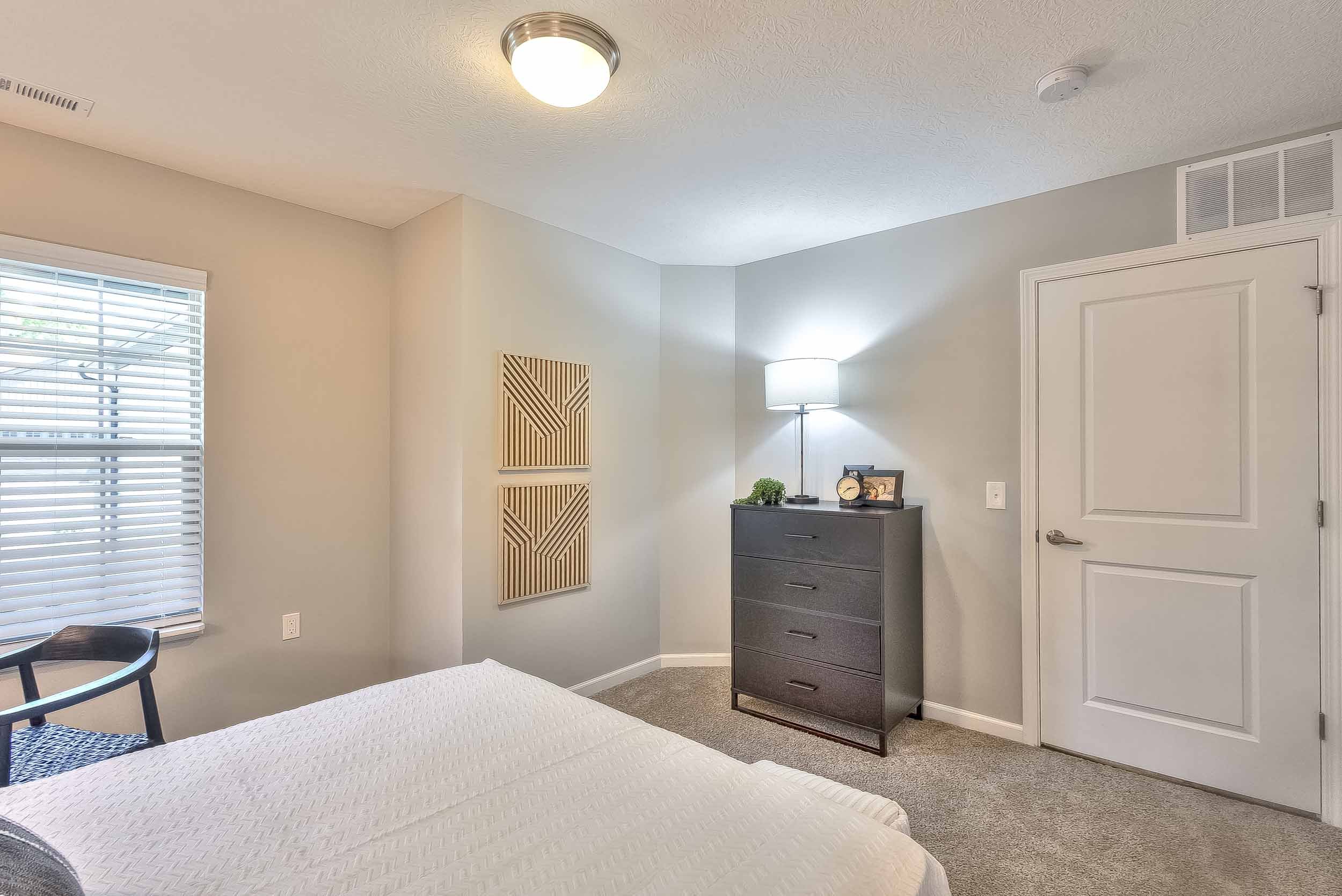 A bedroom with a window to the left, a bed in the foreground, a gray dresser with a lamp on top near the corner, and wall art with geometric patterns on the wall. The room has a beige carpet and light-colored walls.