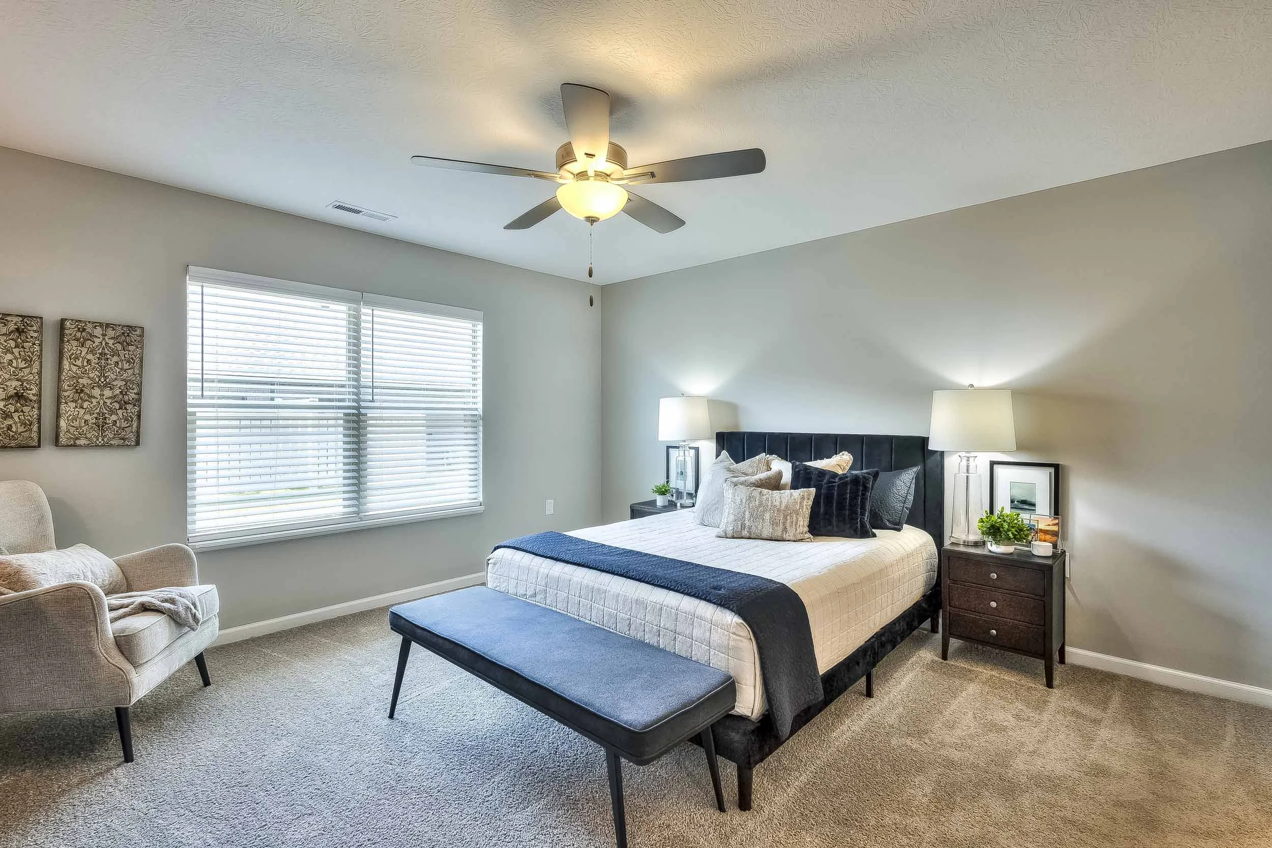 A bedroom with beige carpet, a window with white blinds, a dark upholstered bed with pillows, two matching bedside lamps, a black headboard, a black bench at the foot of the bed, and a beige armchair near the window.