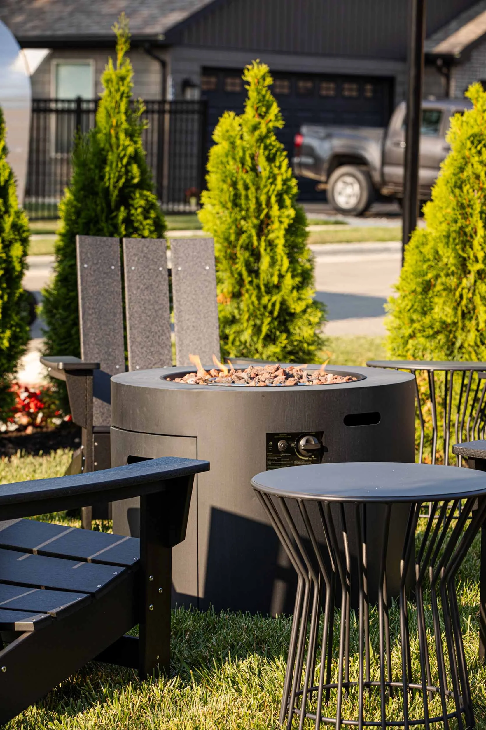 Outdoor patio with fire pit, black chairs, and metal side table, surrounded by tall shrubs and a residential house in the background.