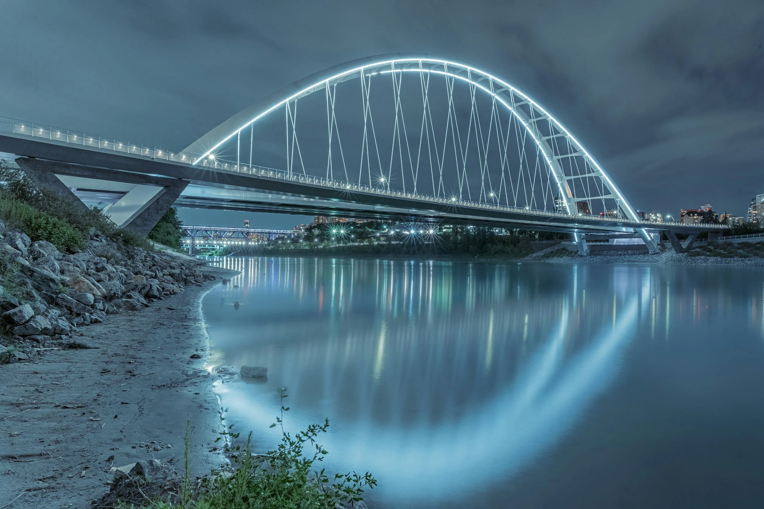 Night view of a modern illuminated arched bridge over a calm river, with city lights in the background and a cloudy sky.