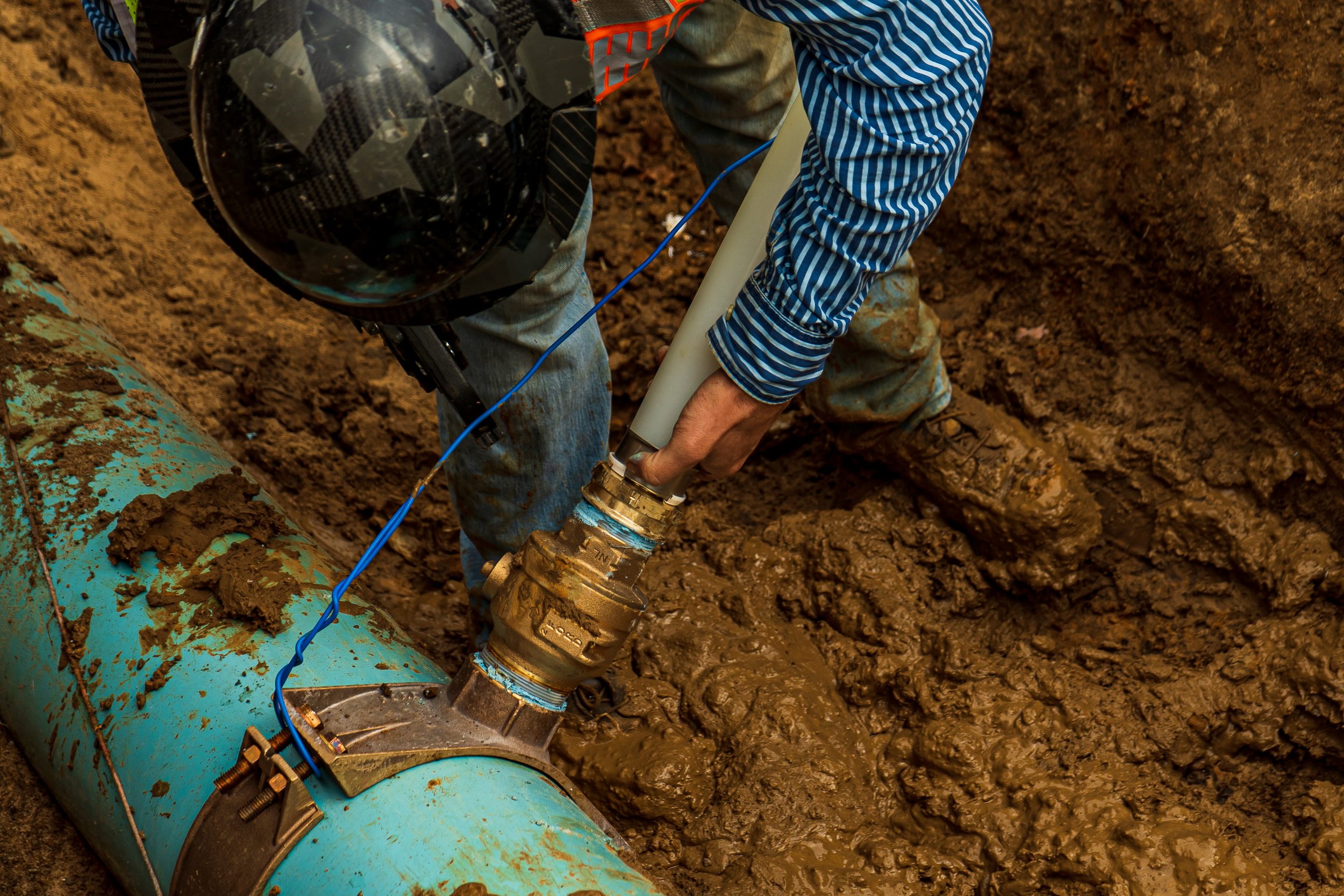 Worker installing a water service connection onto an underground water main inside an excavation trench.