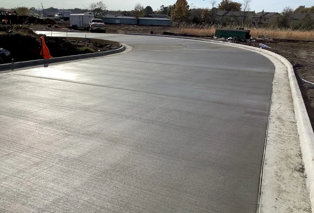 Paved road with curbs shown. Some vehicles and construction equipment in the background.