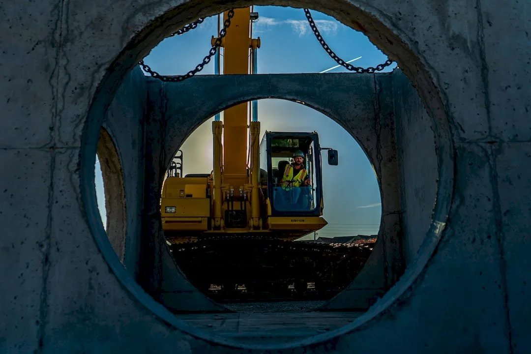View through a concrete pipe showing a construction worker operating a yellow excavator, with the sky in the background.