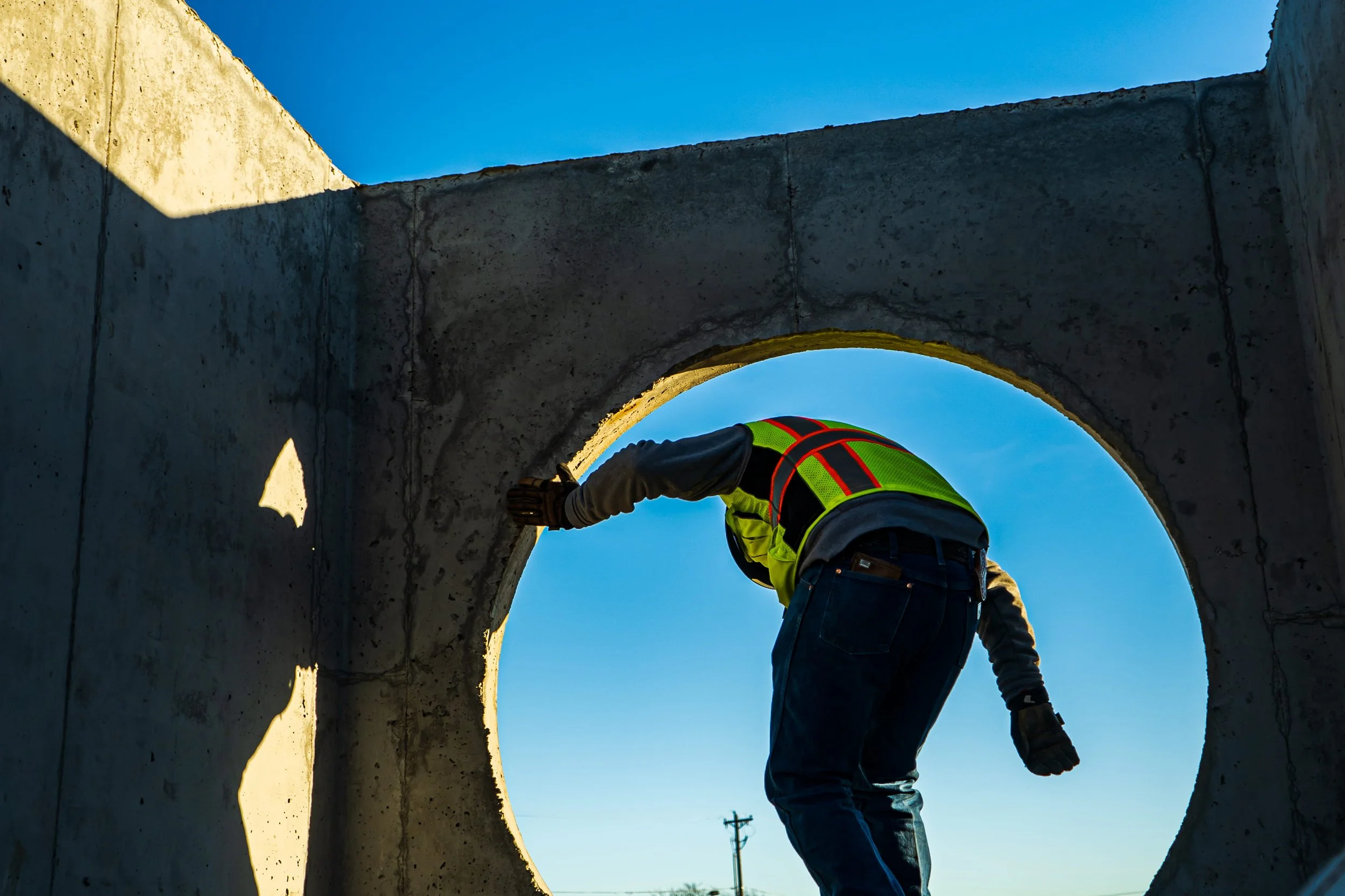 Construction worker entering a precast concrete manhole structure with a circular pipe opening during underground utility installation, viewed from inside the structure against a clear blue sky.
