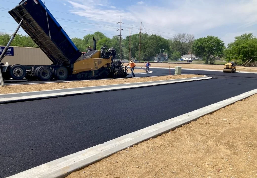 Paved road with curbs shown. Some vehicles and construction equipment in the background.