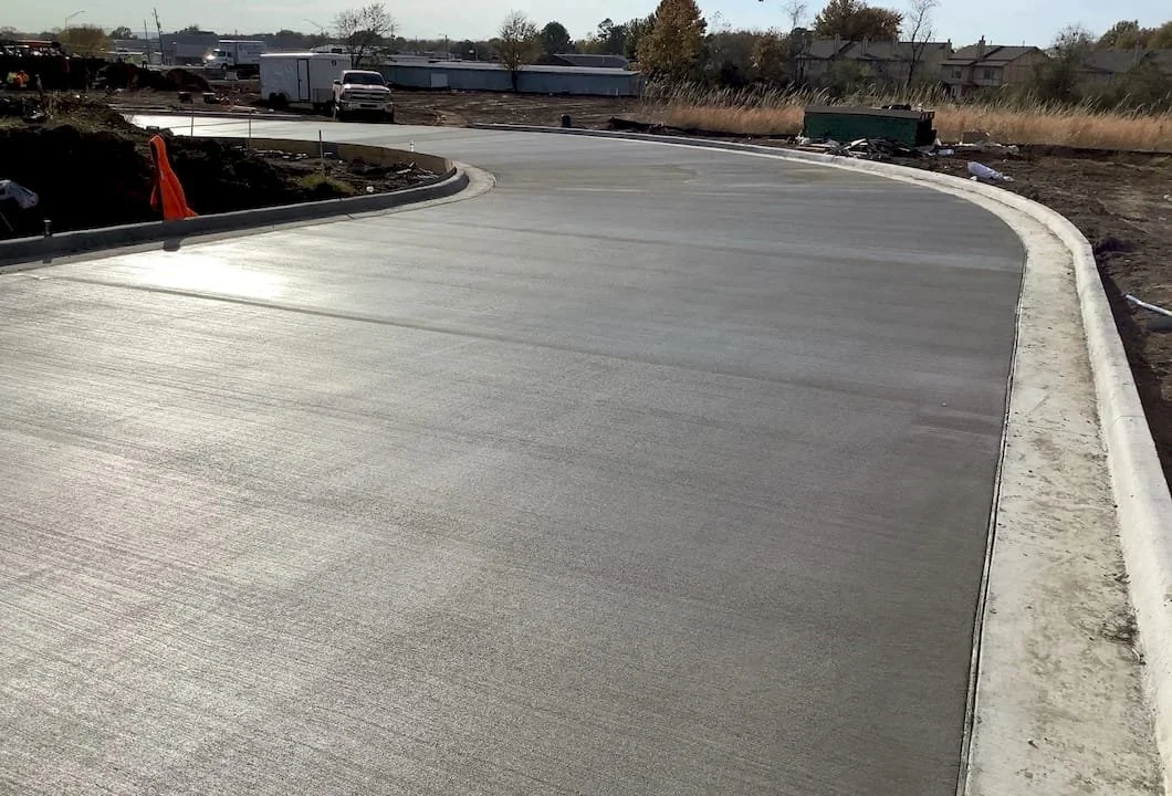 Paved road with curbs shown. Some vehicles and construction equipment in the background.