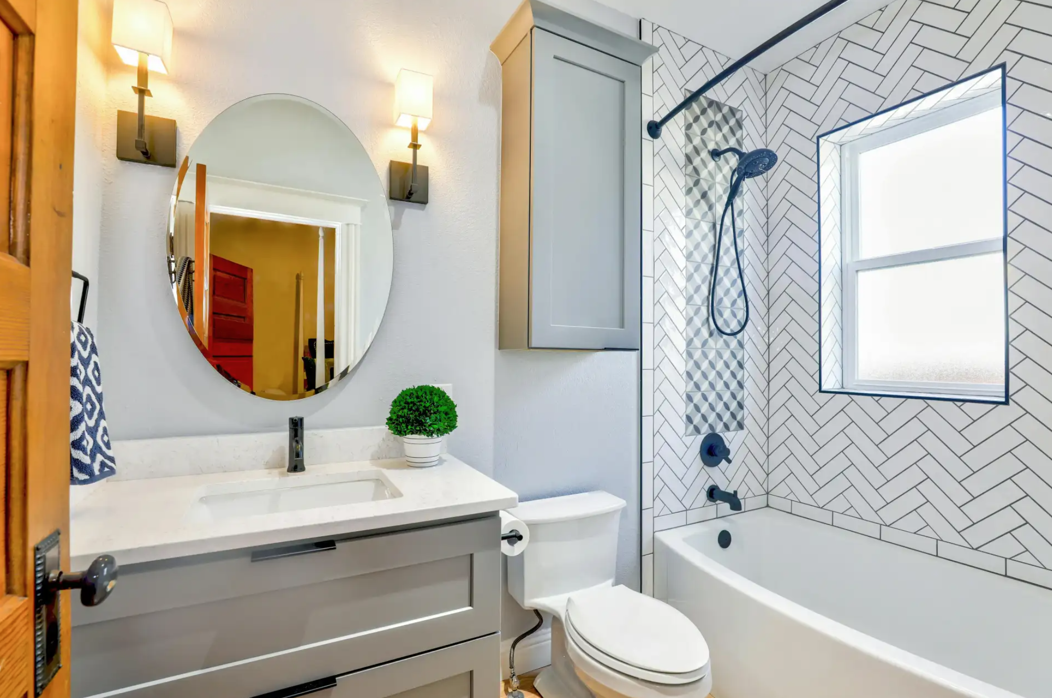 A modern bathroom with a gray vanity, a round mirror, black faucet, small potted plant, wall-mounted light fixtures, a toilet, and a bathtub with a black showerhead and a window in the tiled shower area.