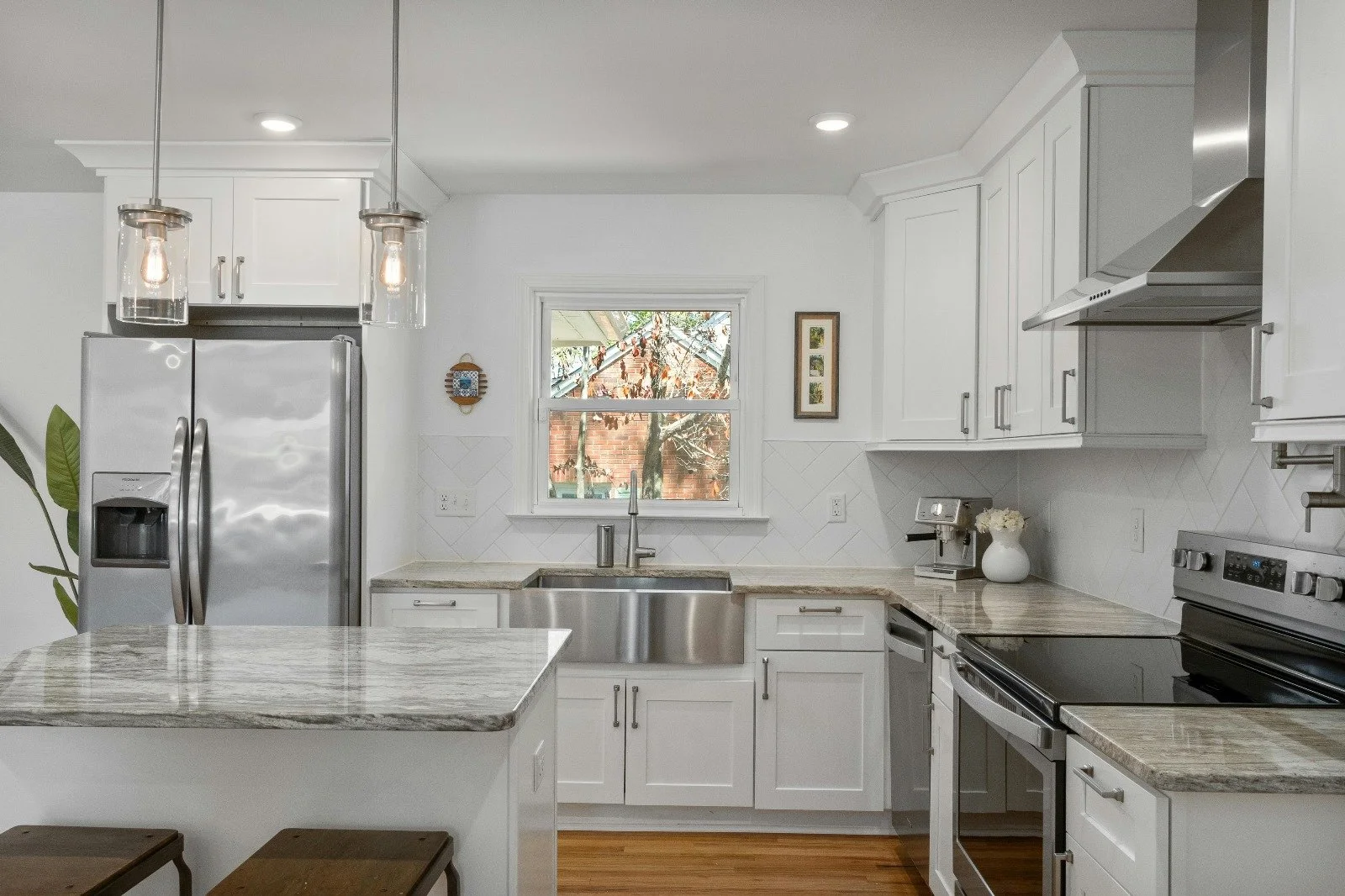 Kitchen with white cabinets, stainless steel refrigerator, oven, and microwave, marble countertops, sink under a window, pendant lights, and hardwood flooring.