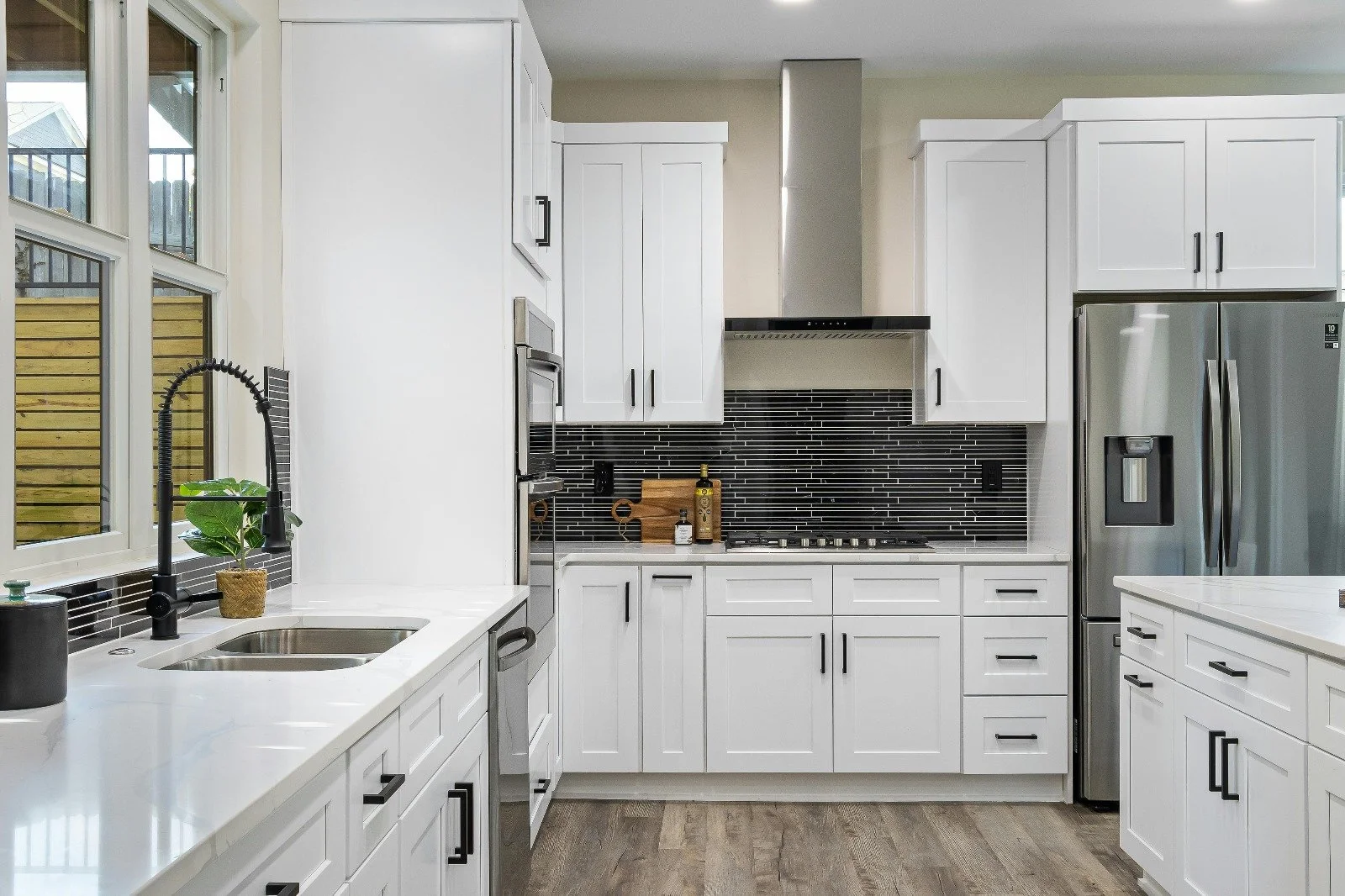 Modern kitchen with white cabinets, black backsplash, stainless steel refrigerator, oven, and sink with black faucet, wooden floor, and window with outside view.