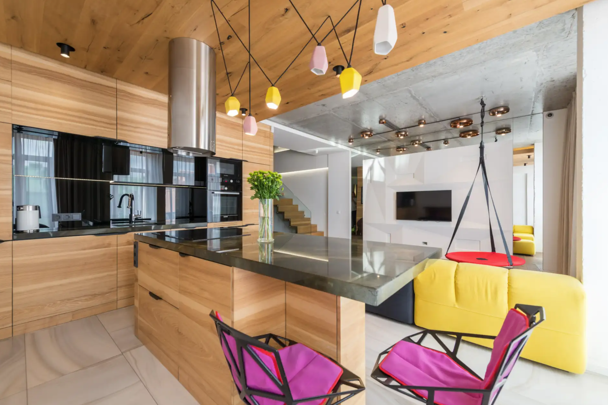 Open-concept kitchen with wooden cabinets, black countertops, and colorful pendant lights. Adjacent living space with yellow couches, a mounted flat-screen TV, and a red swing seat.