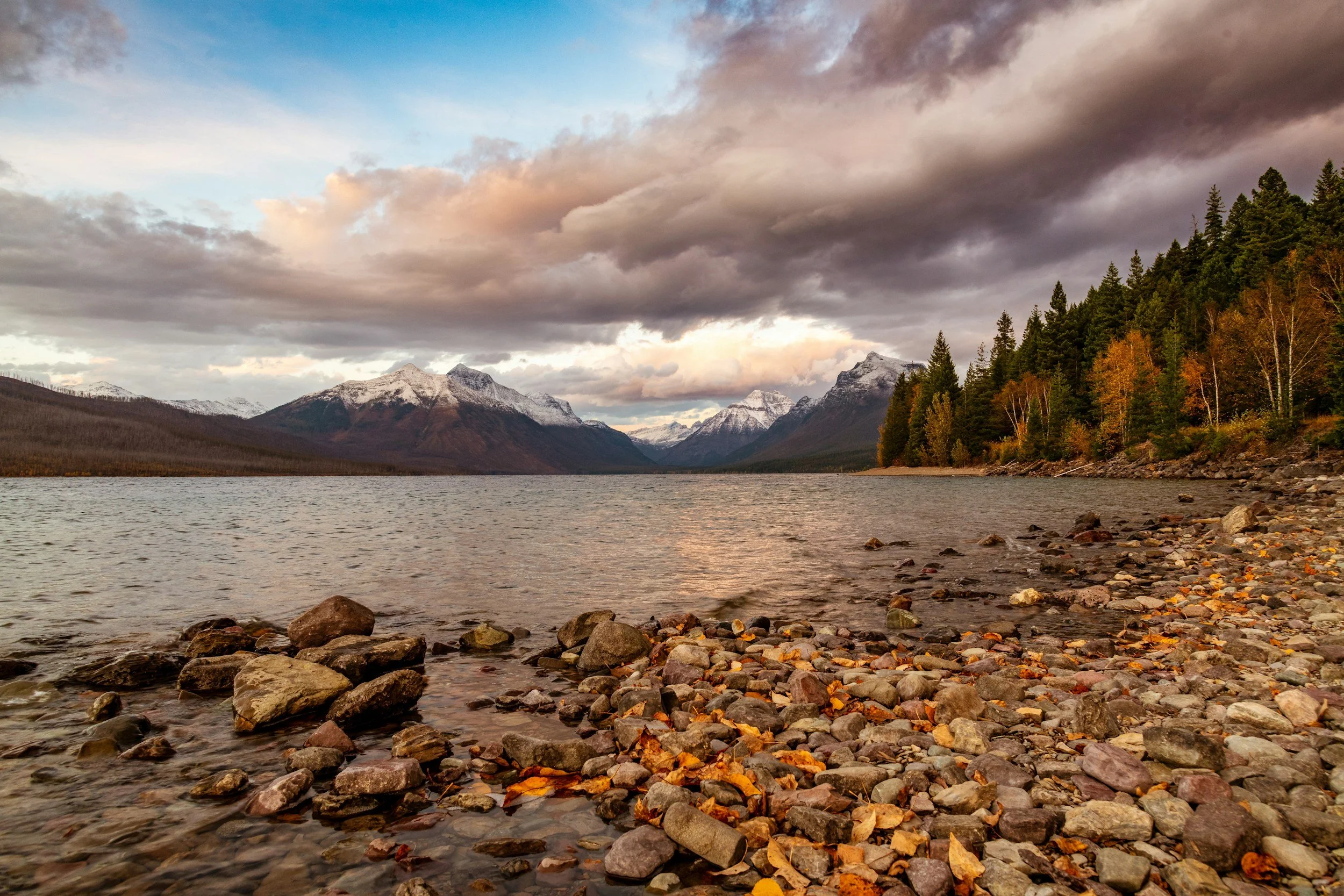 A scenic view of a mountainous landscape with snow-capped peaks, a lake in the foreground, and a forest with changing autumn leaves on the right side, all under a sky filled with dark and light clouds.