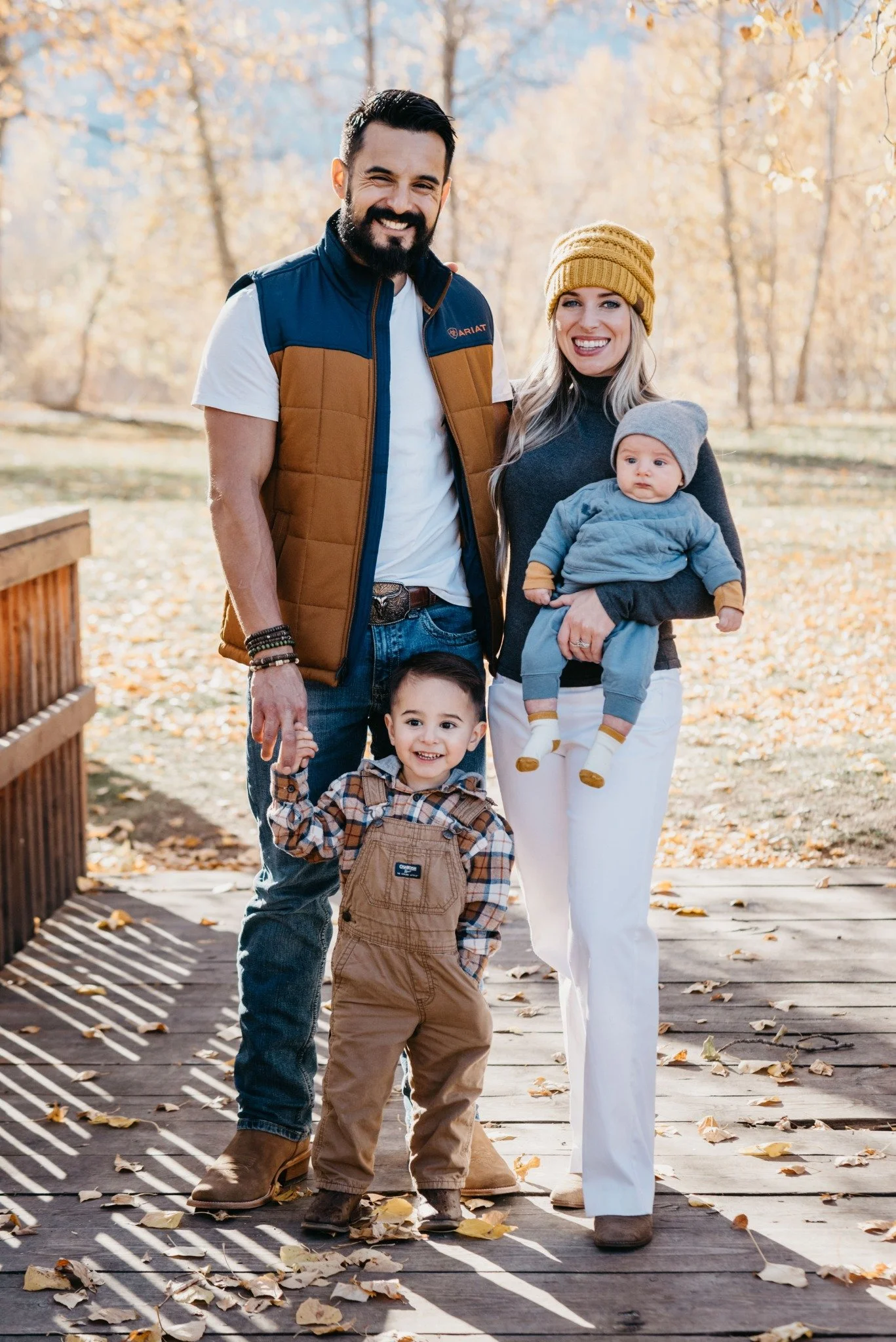 A family of four standing on a wooden bridge in autumn, smiling at the camera. The father has dark hair and a beard, wearing a brown and navy vest and jeans. The mother has long blonde hair, wearing a mustard yellow beanie, dark top, and white pants. She is holding a baby dressed in gray and blue with a gray beanie. A young boy with dark hair, wearing a plaid shirt and brown overalls, stands beside them holding his father's hand.