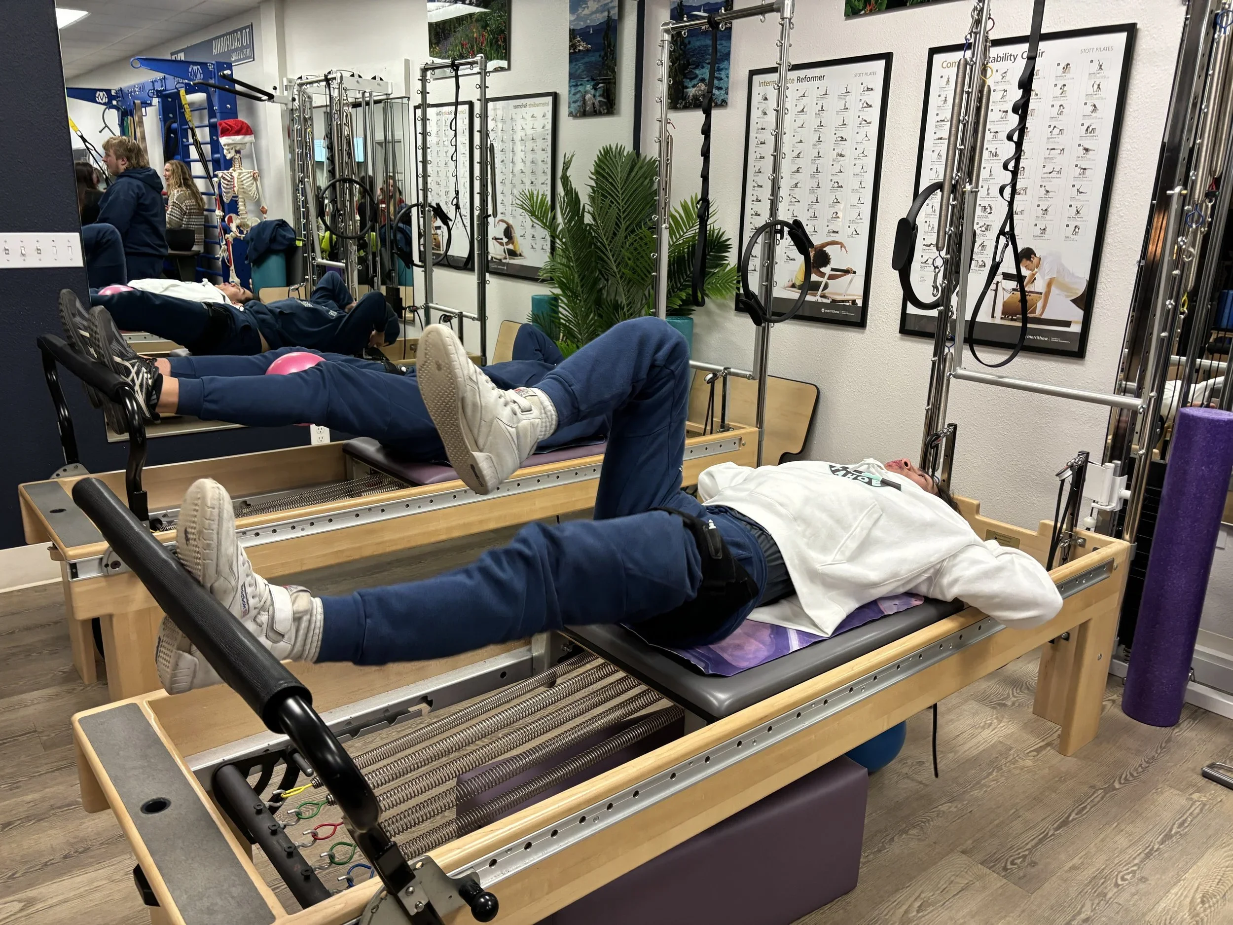 Two people lying on reformer Pilates machines in a fitness studio, with various equipment and posters on the wall.