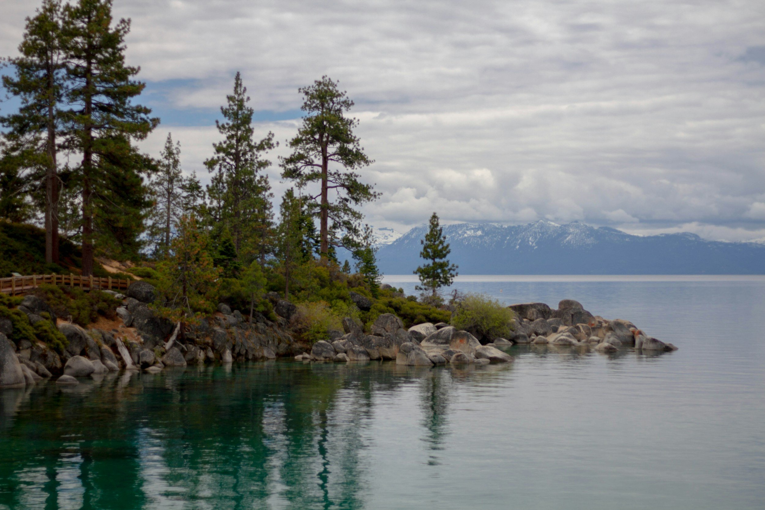 A peaceful lakeside scene in Tahoe with a rocky shoreline, green trees, calm water, and snow-capped mountains in the background under a cloudy sky.