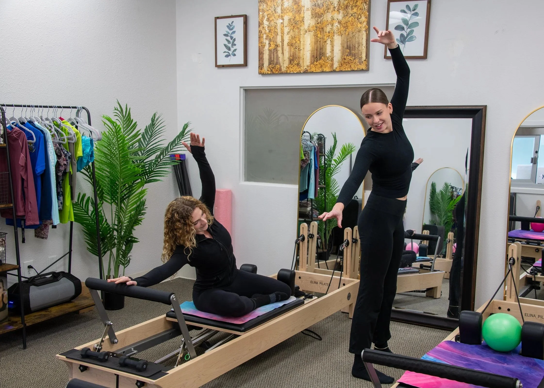 A woman doing Pilates on a reformer machine in a fitness studio while a trainer guides her. The studio has large mirrors, framed botanical artwork, exercise equipment, and plants.