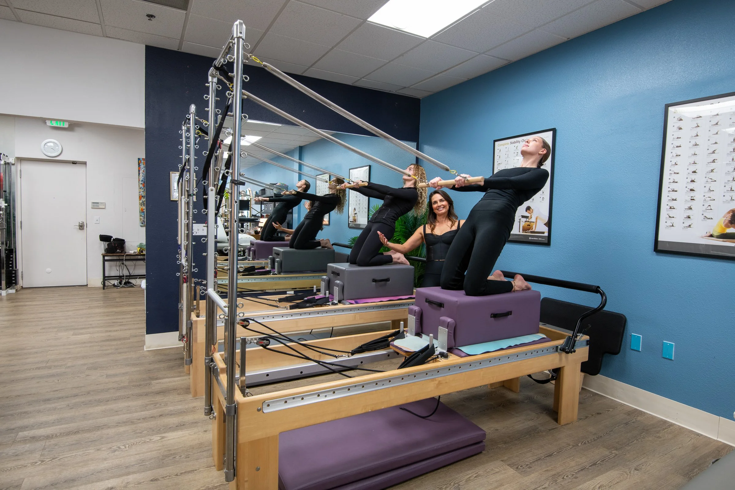 Four women participating in a Pilates Reformer class in a fitness studio, guided by an instructor.