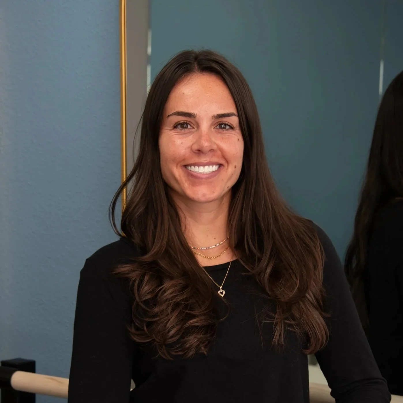 A woman with long brown hair, smiling, wearing a black top and layered necklaces, standing indoors against a teal wall.