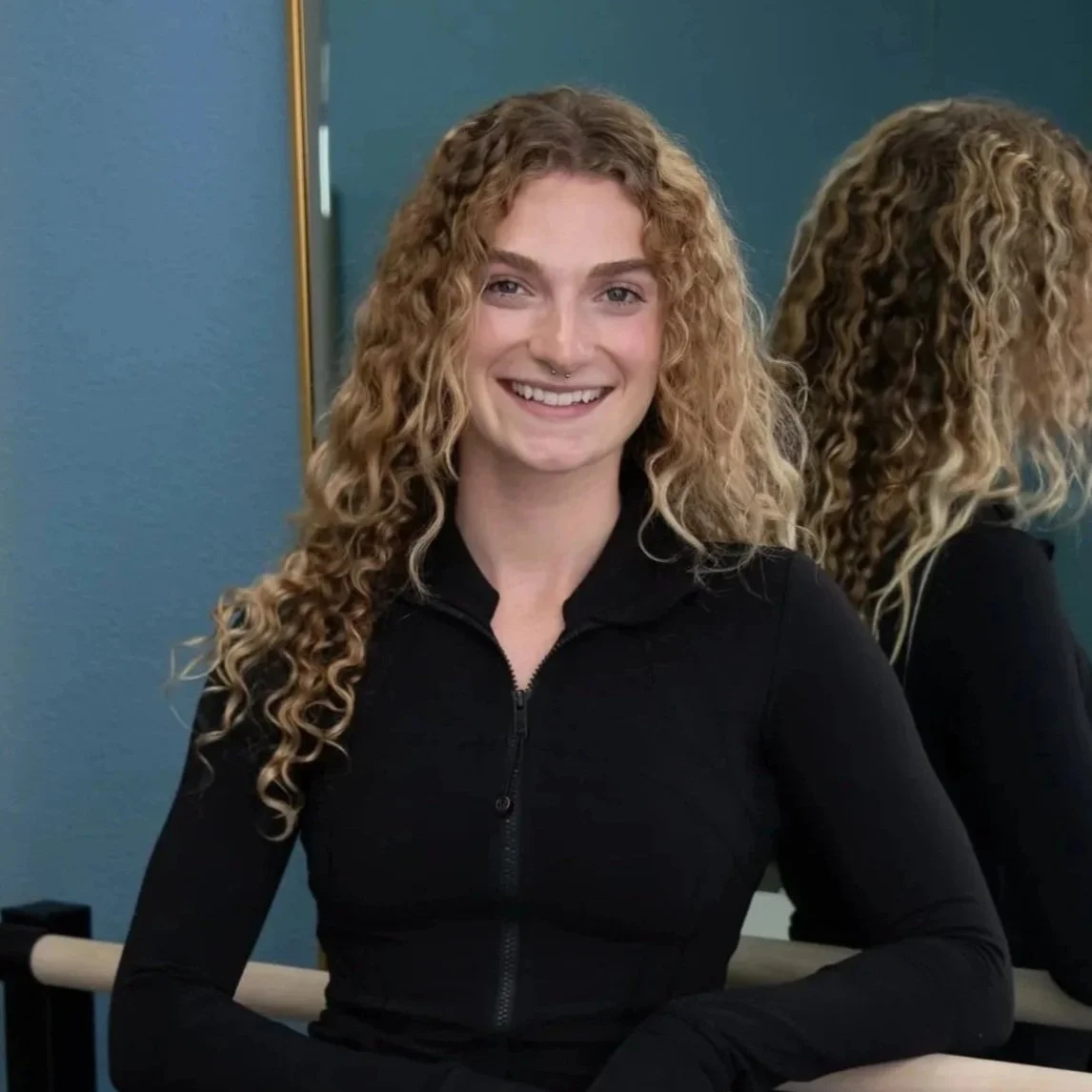 A woman with curly blonde hair and a nose piercing smiling at the camera, standing in front of a mirror in a room with blue walls.