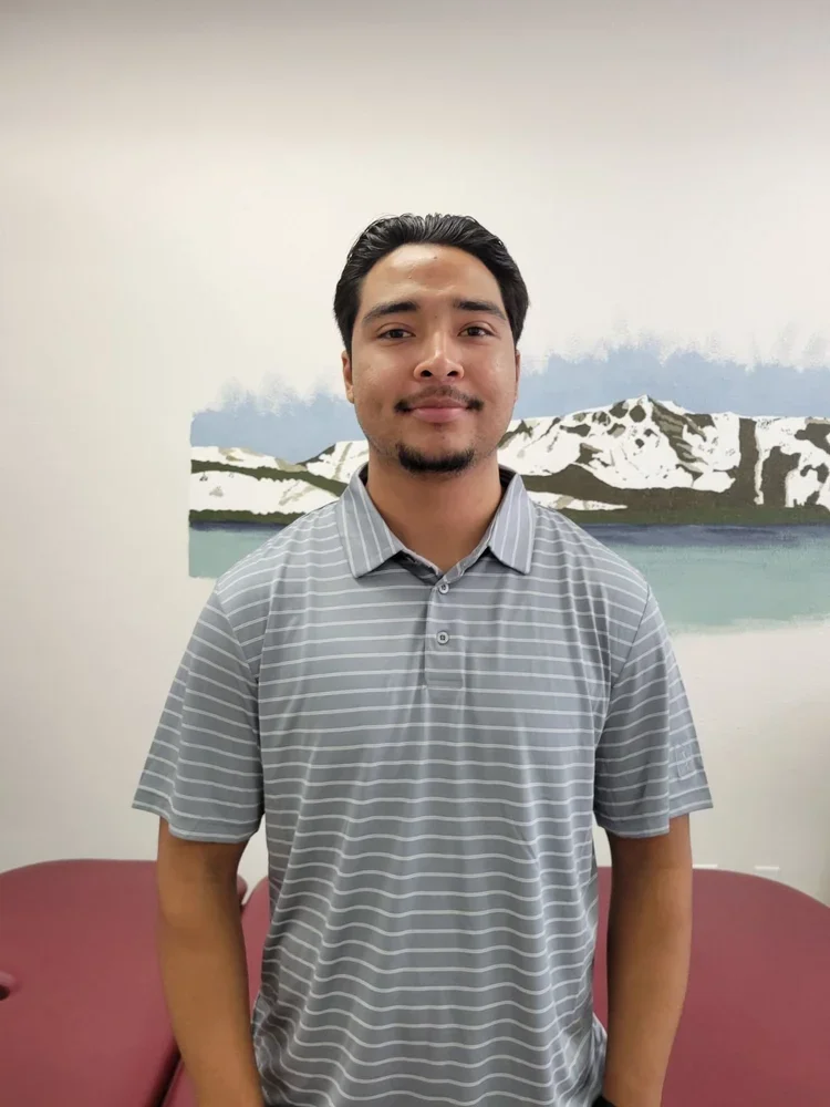 A young man standing in front of a painted landscape of mountains and water, wearing a gray striped polo shirt.