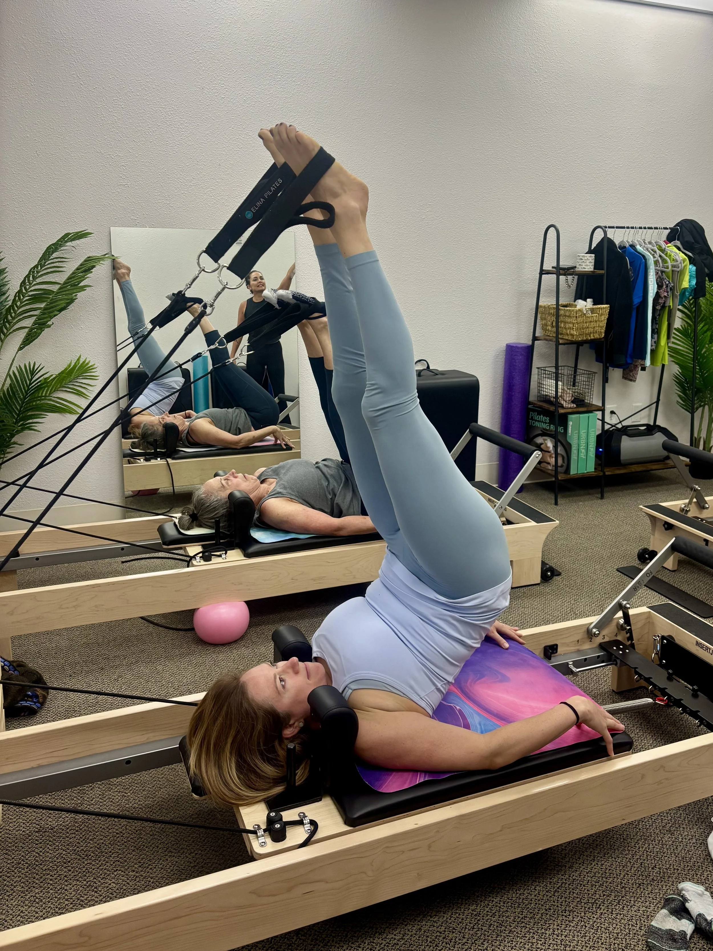 A woman lying on a Pilates reformer machine with her legs raised and strapped in a Pilates ring, practicing Pilates exercises. In the background, she can see her reflection in a mirror, along with another person exercising.