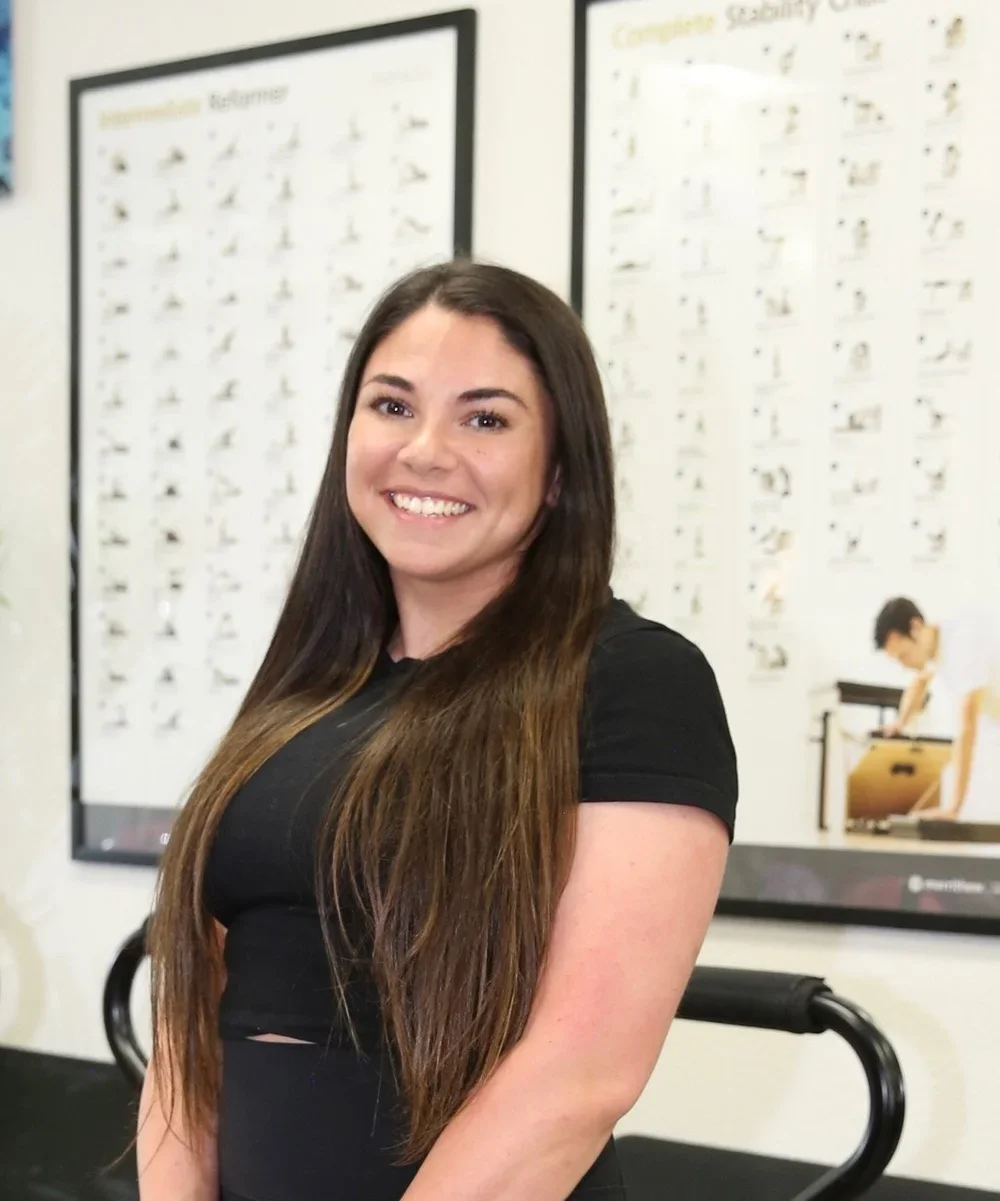 A young woman with long brown hair and a black shirt smiling indoors, with posters of exercise movements on the wall behind her.