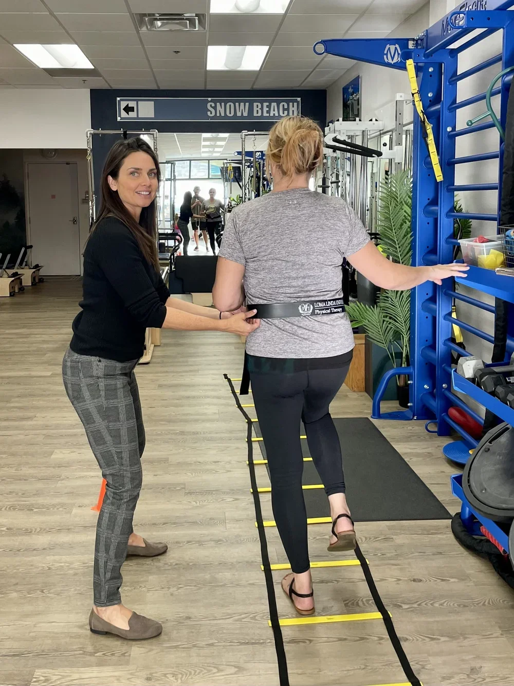 A woman is participating in a physical therapy session with a therapist at a gym, walking on a yellow and black agility ladder on the floor in front of a wall-mounted blue treatment strap.
