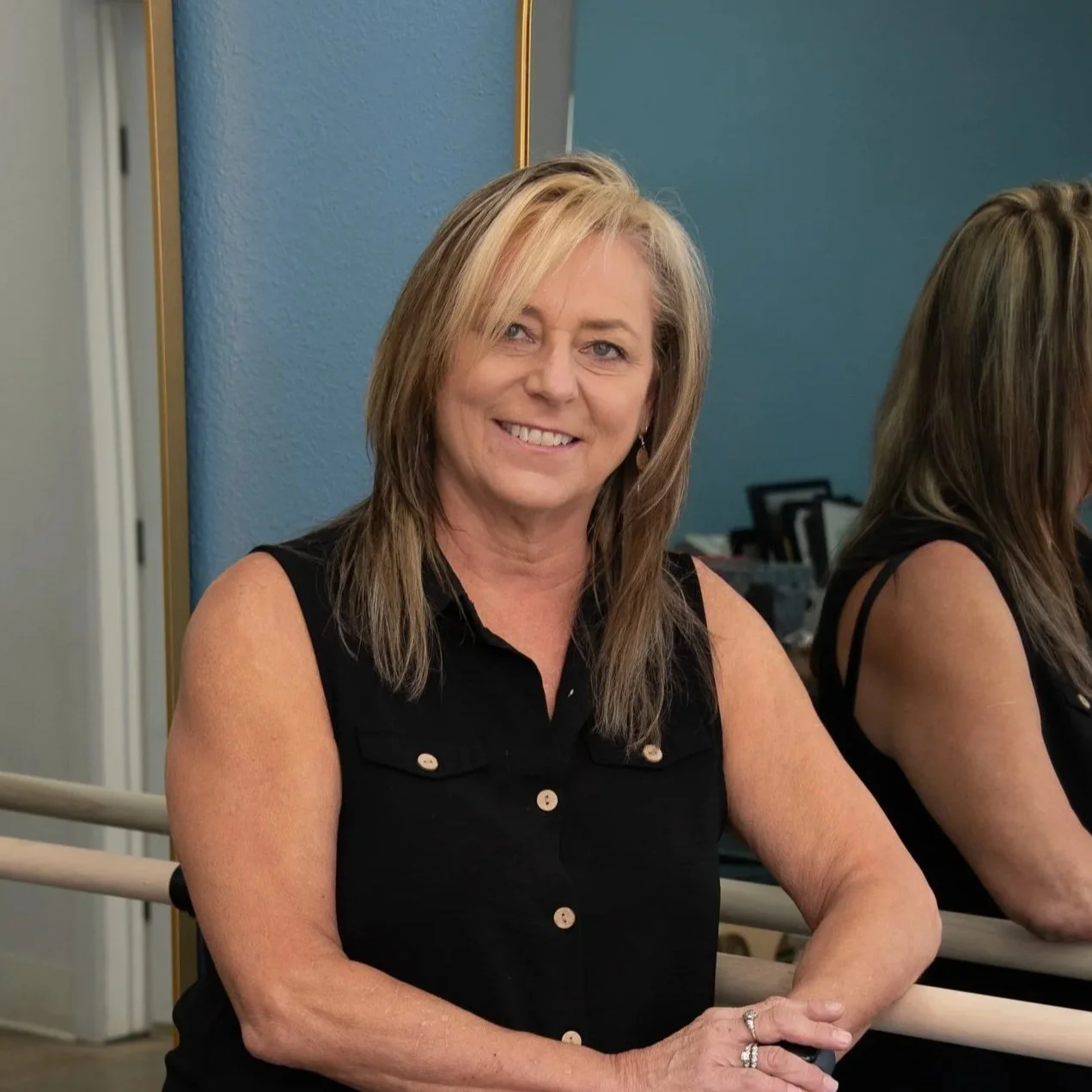 A woman with blonde hair in a sleeveless black top, smiling, sitting at a mirror in a room with a blue wall.
