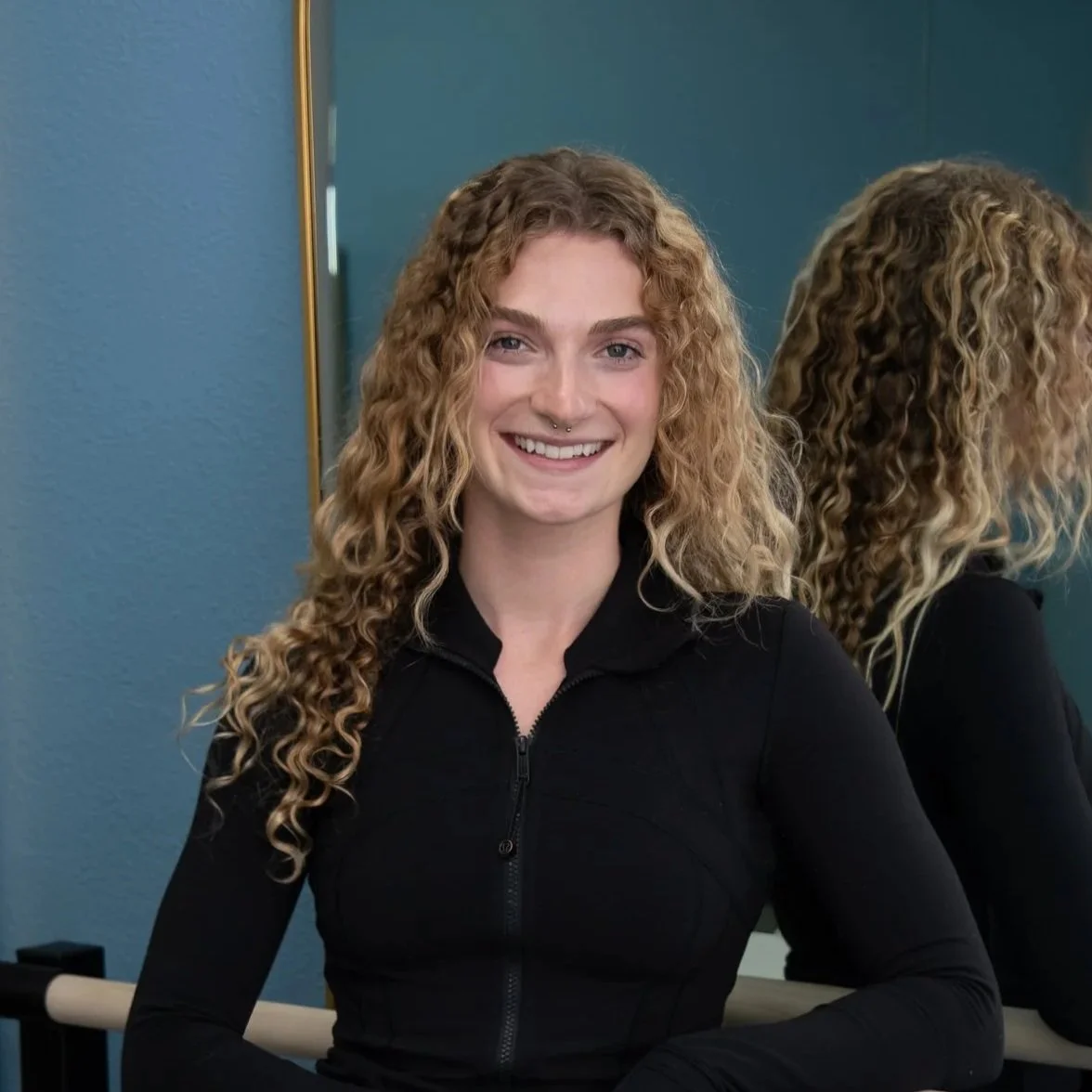 A woman with long, curly blonde hair smiling at the camera, standing in front of a mirror, wearing a black zip-up shirt.