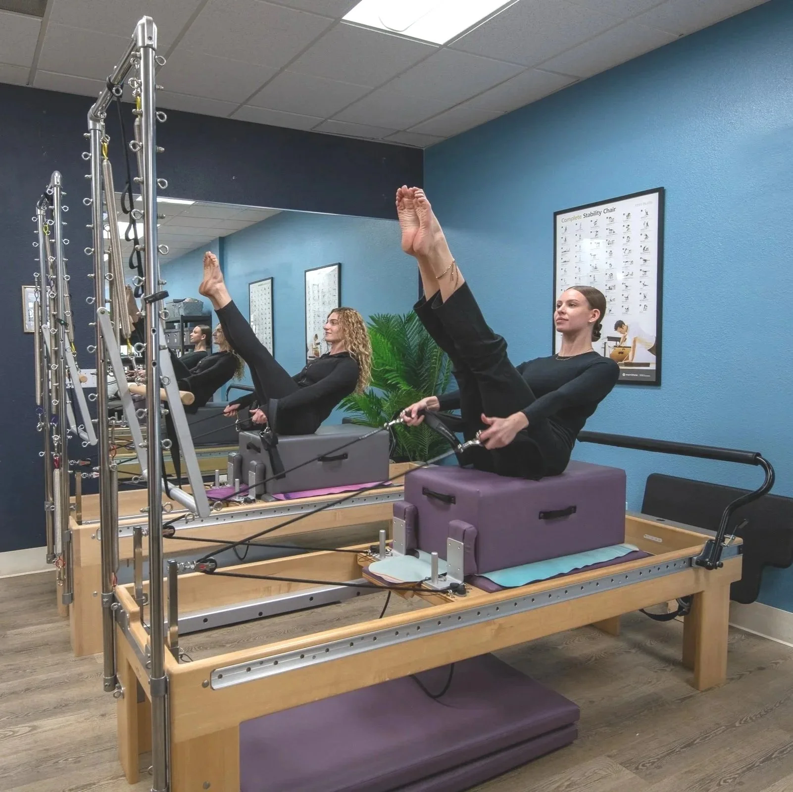 Two women in black workout clothes lying on reformer Pilates machines, performing leg lifts with straps attached to their ankles in a Pilates studio with blue walls and a mirror.