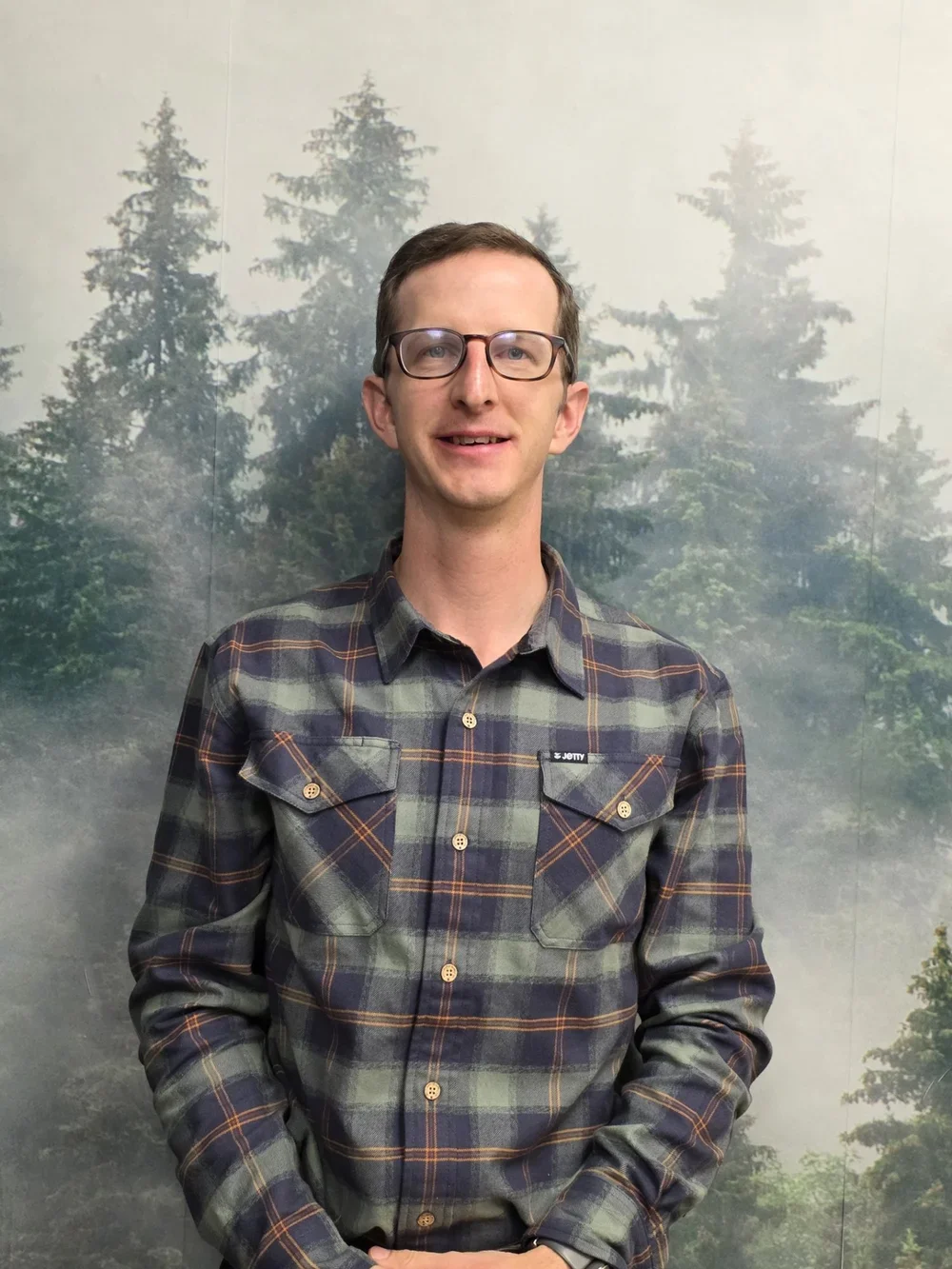 A man with glasses and a plaid shirt standing in front of a backdrop of evergreen trees.