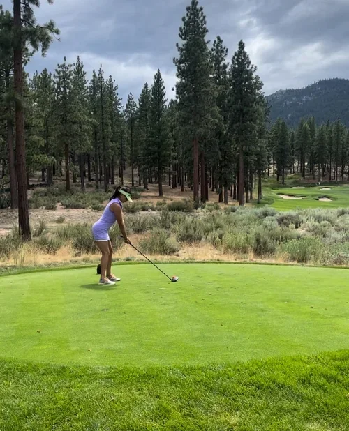 A woman dressed in white is preparing to tee off on a golf course surrounded by pine trees and hills under cloudy skies.