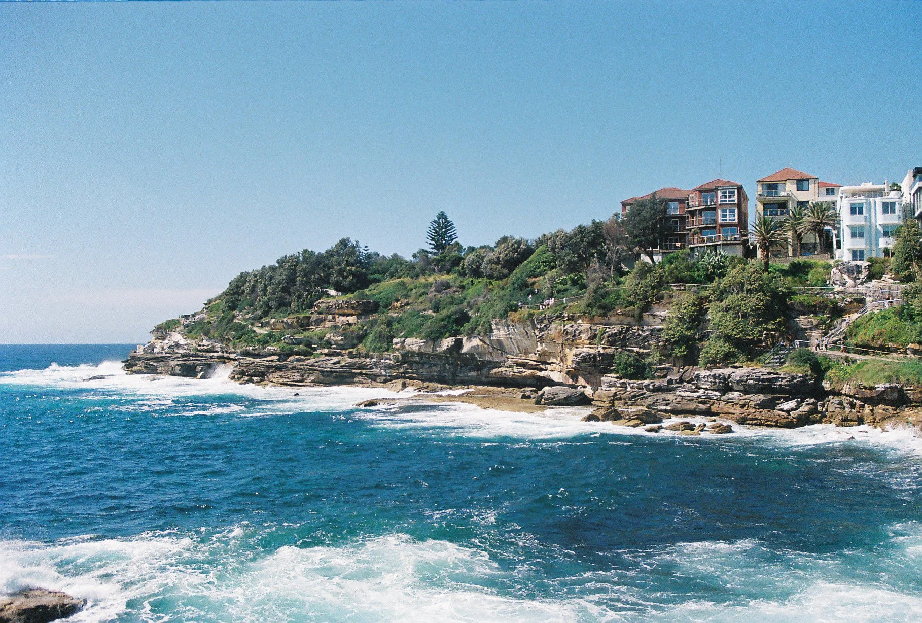Cliffside houses overlooking the ocean with clear blue water and waves hitting the rocks.