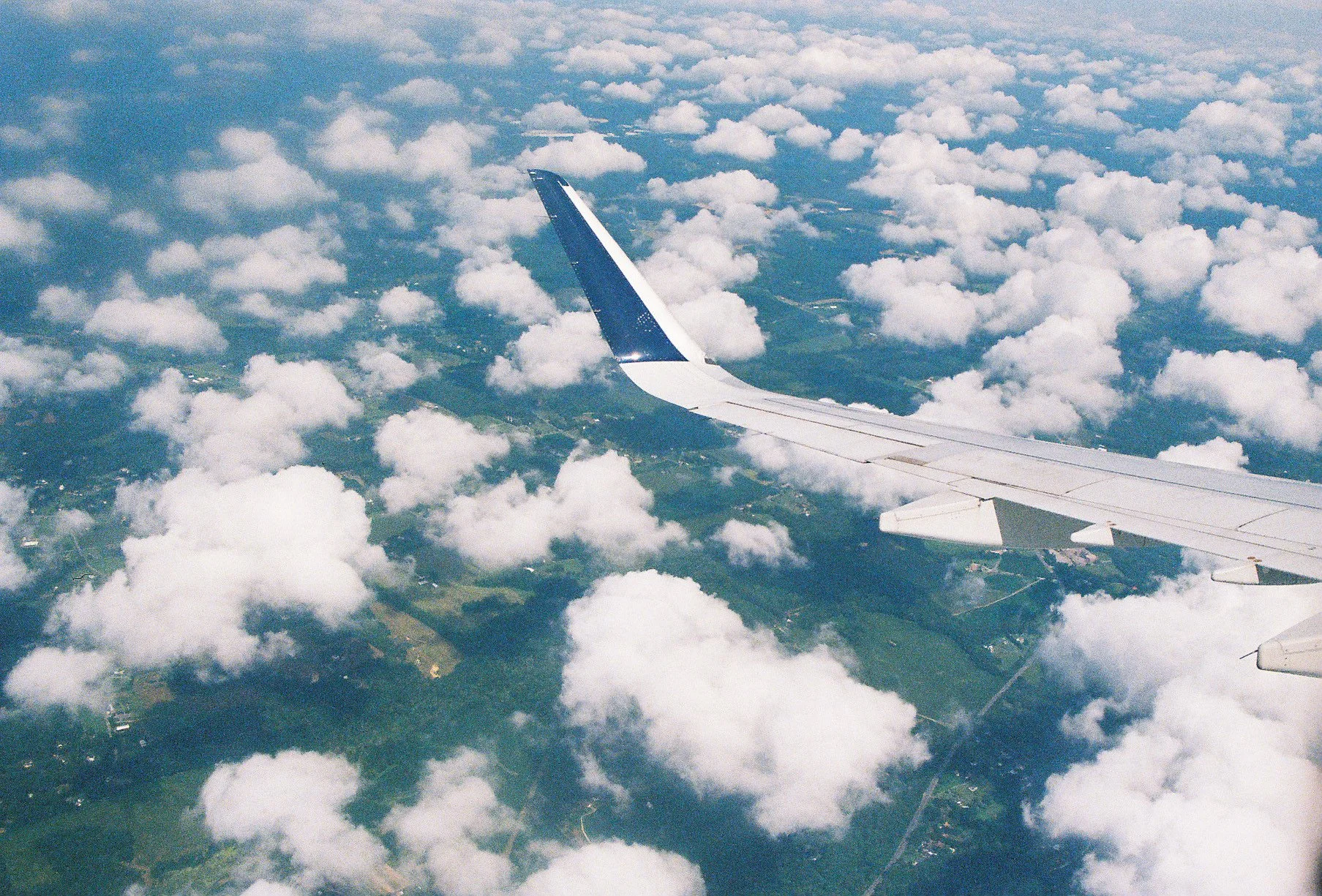 View from airplane window showing the wing and a landscape with green fields and scattered clouds below.