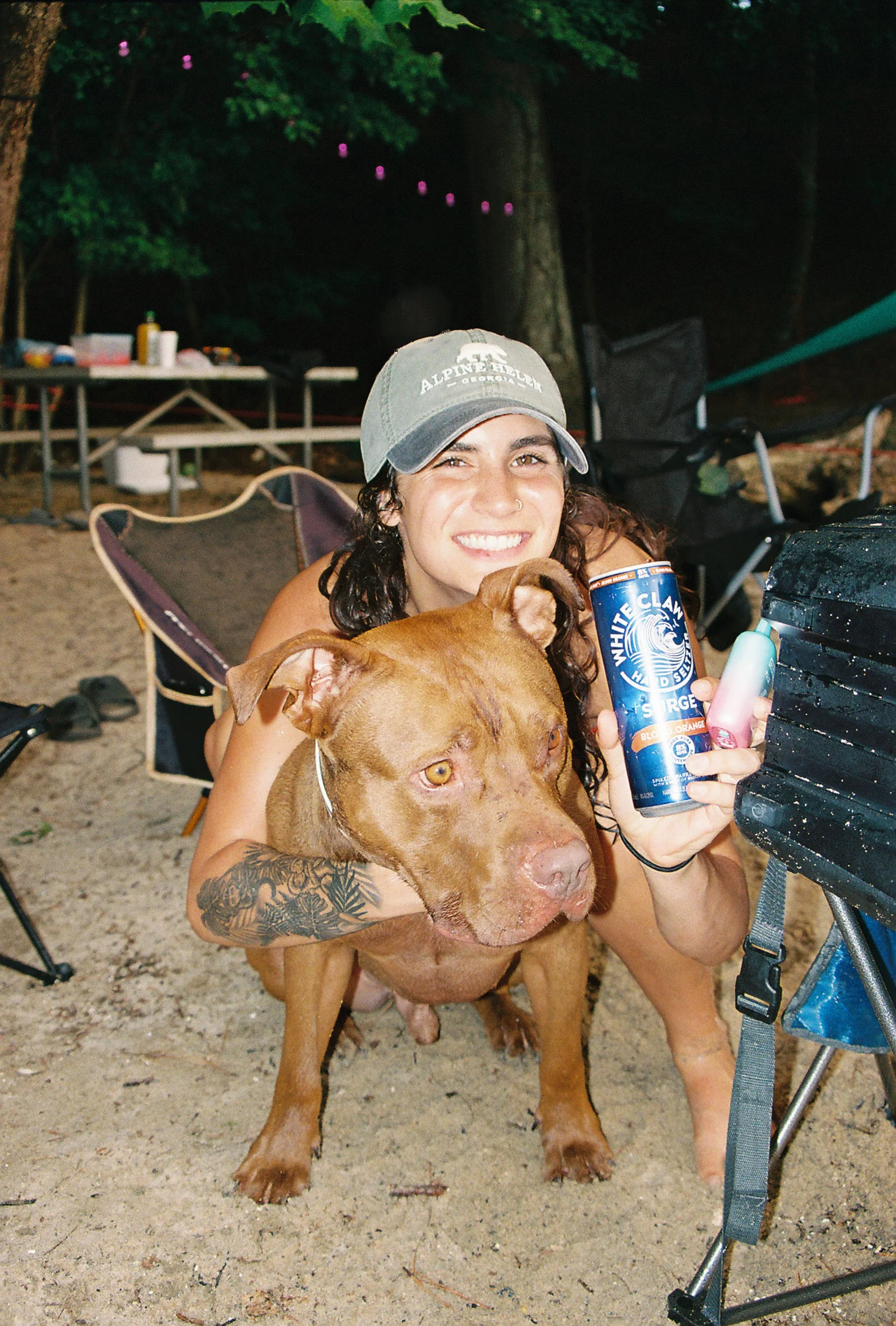 A smiling woman with curly dark hair, wearing a gray cap and holding a can of White Claw hard seltzer, is hugging a large brown dog with a tattoos on its front leg at an outdoor nighttime gathering.