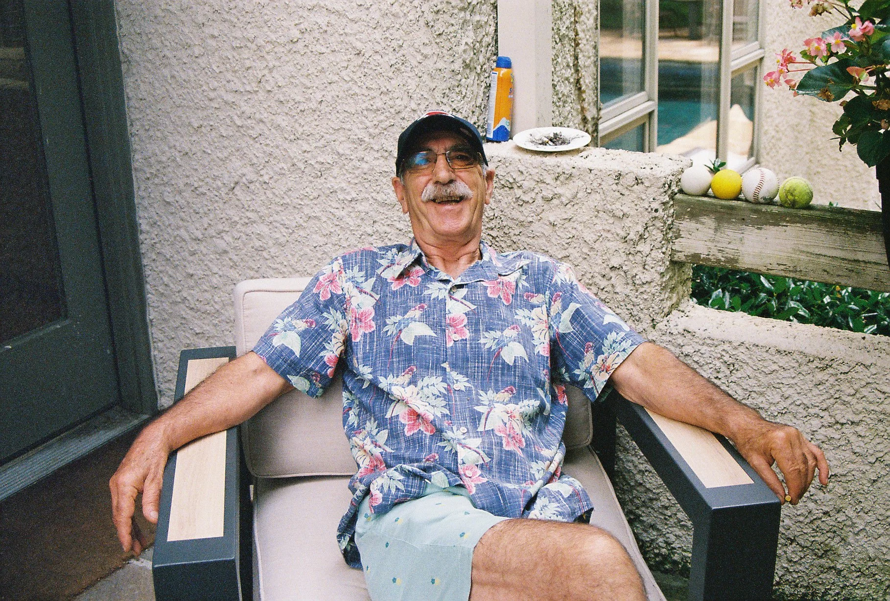 An older man with a mustache and glasses, wearing a tropical floral shirt, shorts, and a cap, sitting relaxed on outdoor patio furniture, smiling, with decorative objects like baseballs, tennis balls, and a lemon on a ledge behind him.