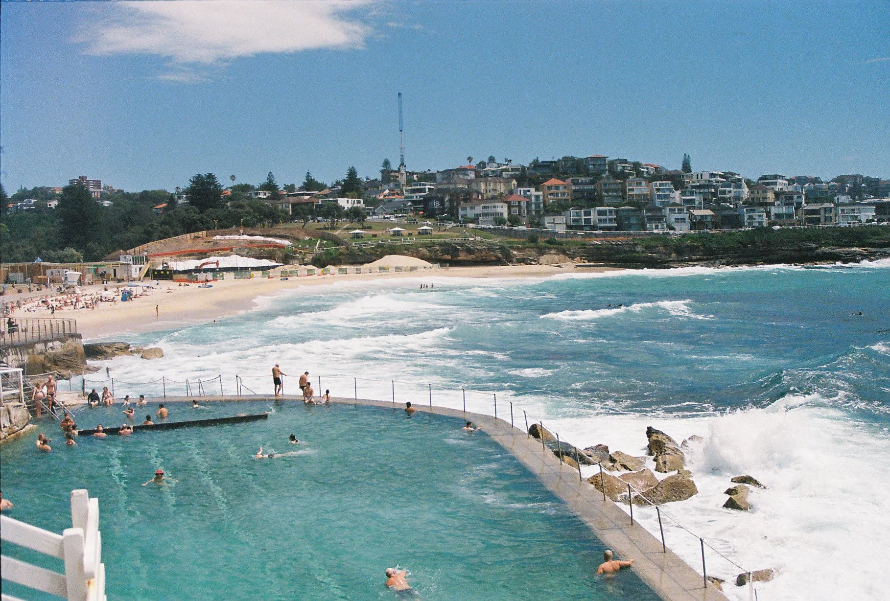 People swimming and relaxing in an ocean pool with waves crashing nearby, beach and residential houses in the background under a blue sky.