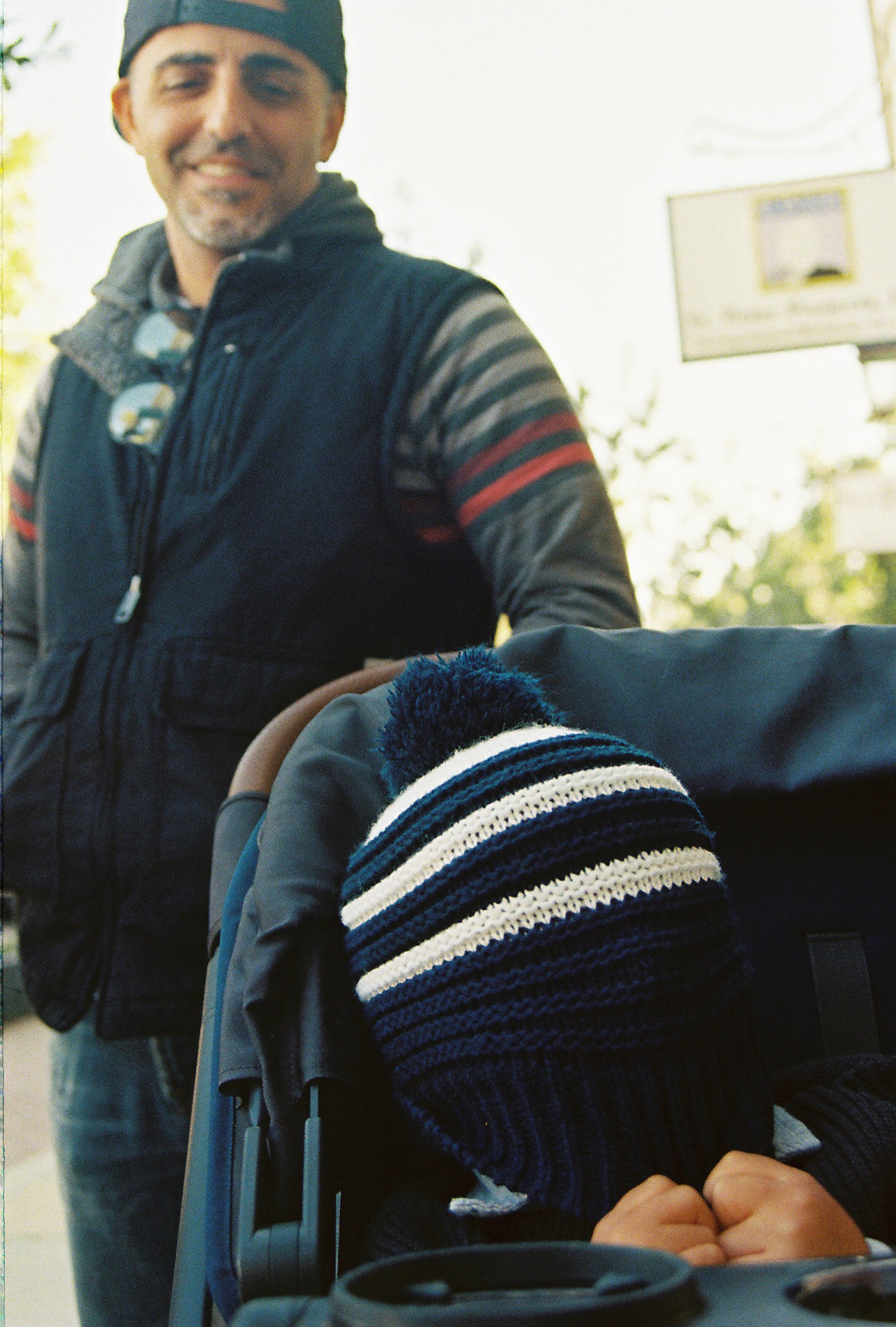 A man smiling at a camera in an outdoor setting, with a child in a stroller in front of him, wearing a striped knit hat with a pom-pom.