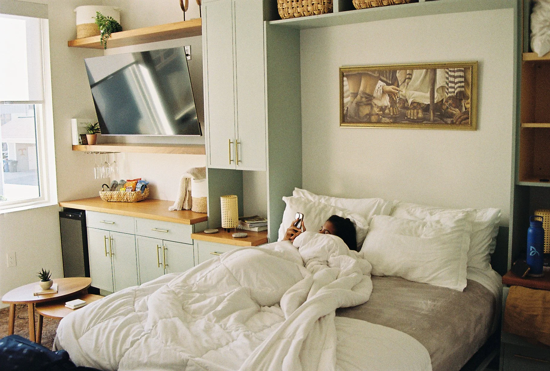 A woman lying in bed using a smartphone in a cozy bedroom with light-colored walls, white bedding, a framed artwork, a wooden nightstand with a blue water bottle, and a built-in shelf with baskets and decorative items.