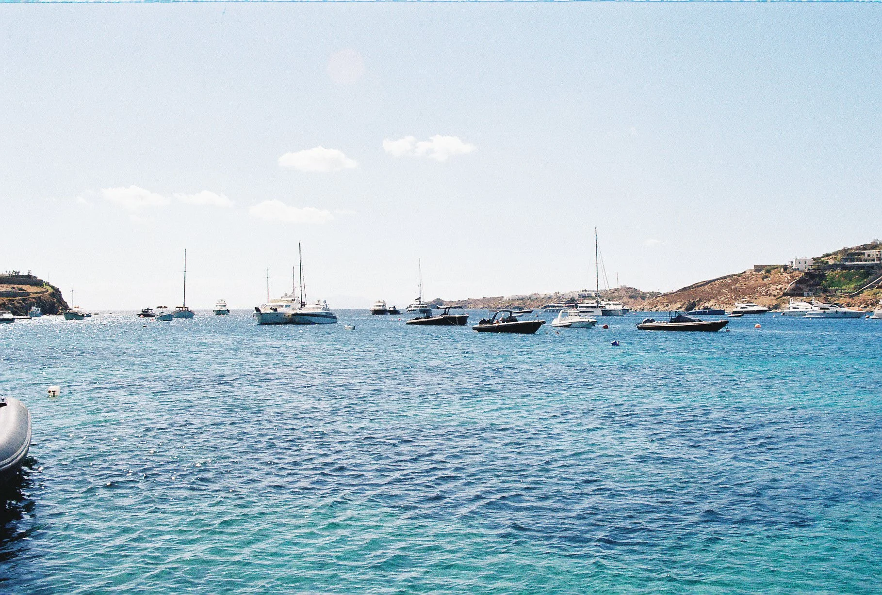 Calm marina with boats and sailboats anchored in bright blue water, hillside with residential buildings in the background, and a partly cloudy sky.