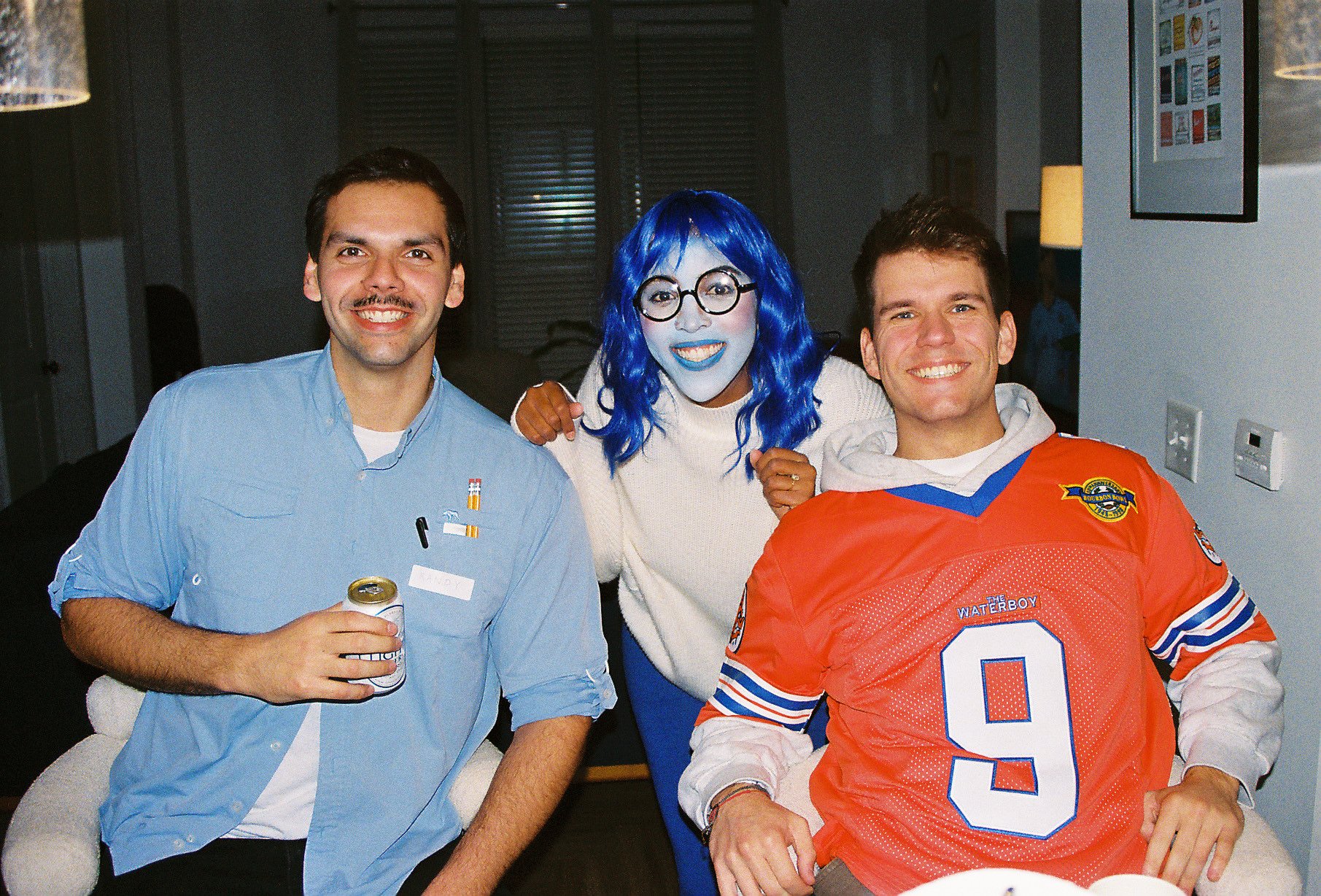 Three people smiling and posing for the camera in a room. The person on the left is a man with mustache and dark hair, wearing a light blue shirt, holding a soda can. The person in the middle is a woman with blue hair, glasses, and painted blue face,