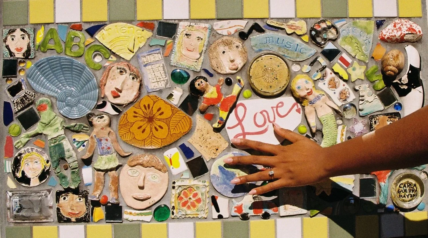 A colorful mosaic collage with various small, hand-crafted figurines, drawings, and objects including faces, animals, and symbols, with a hand pointing to a piece that says "Love".